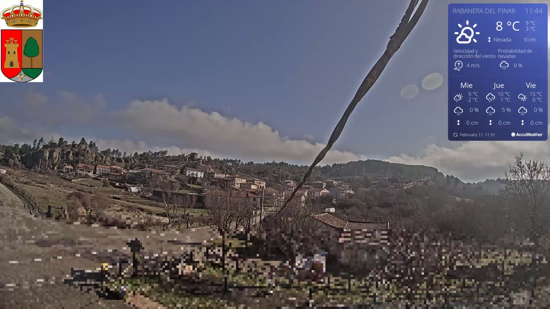 A partly cloudy day reveals a small village nestled in a hillside valley, with brown and tan buildings visible against a backdrop of trees and low hills.