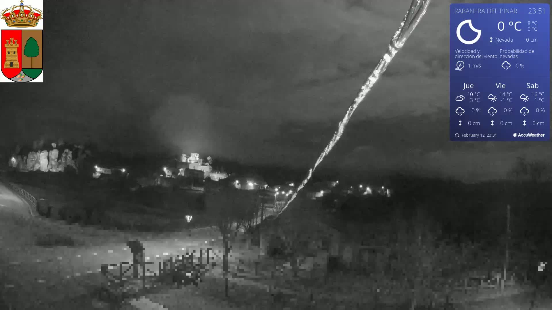 A nighttime, low-light view of a small town under a dark sky with a bright streak across it, at 0°C with a 1 m/s wind and no snow.