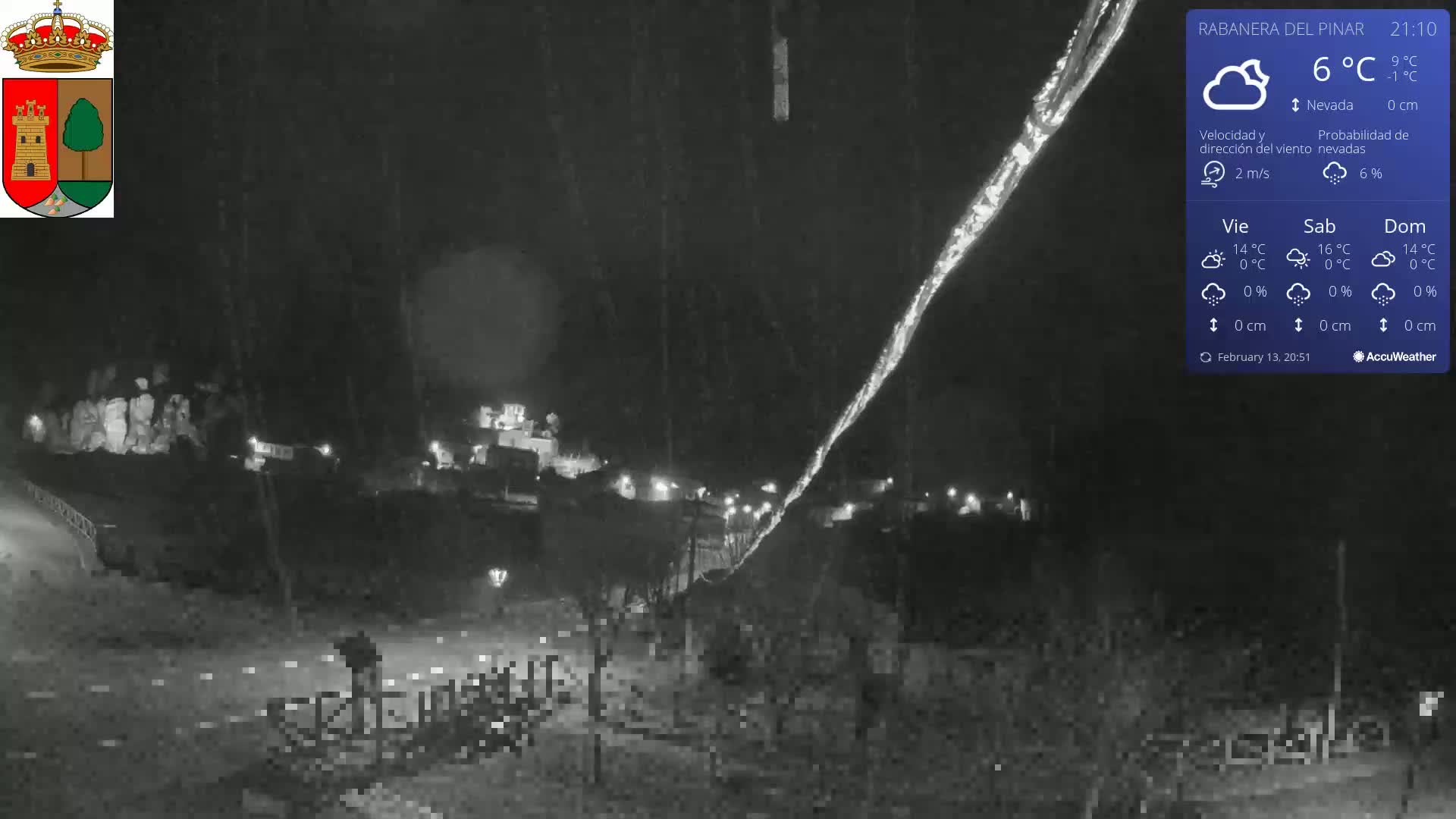 A nighttime, snow-covered scene shows a small village illuminated with faint lights under a lightly snowing sky.