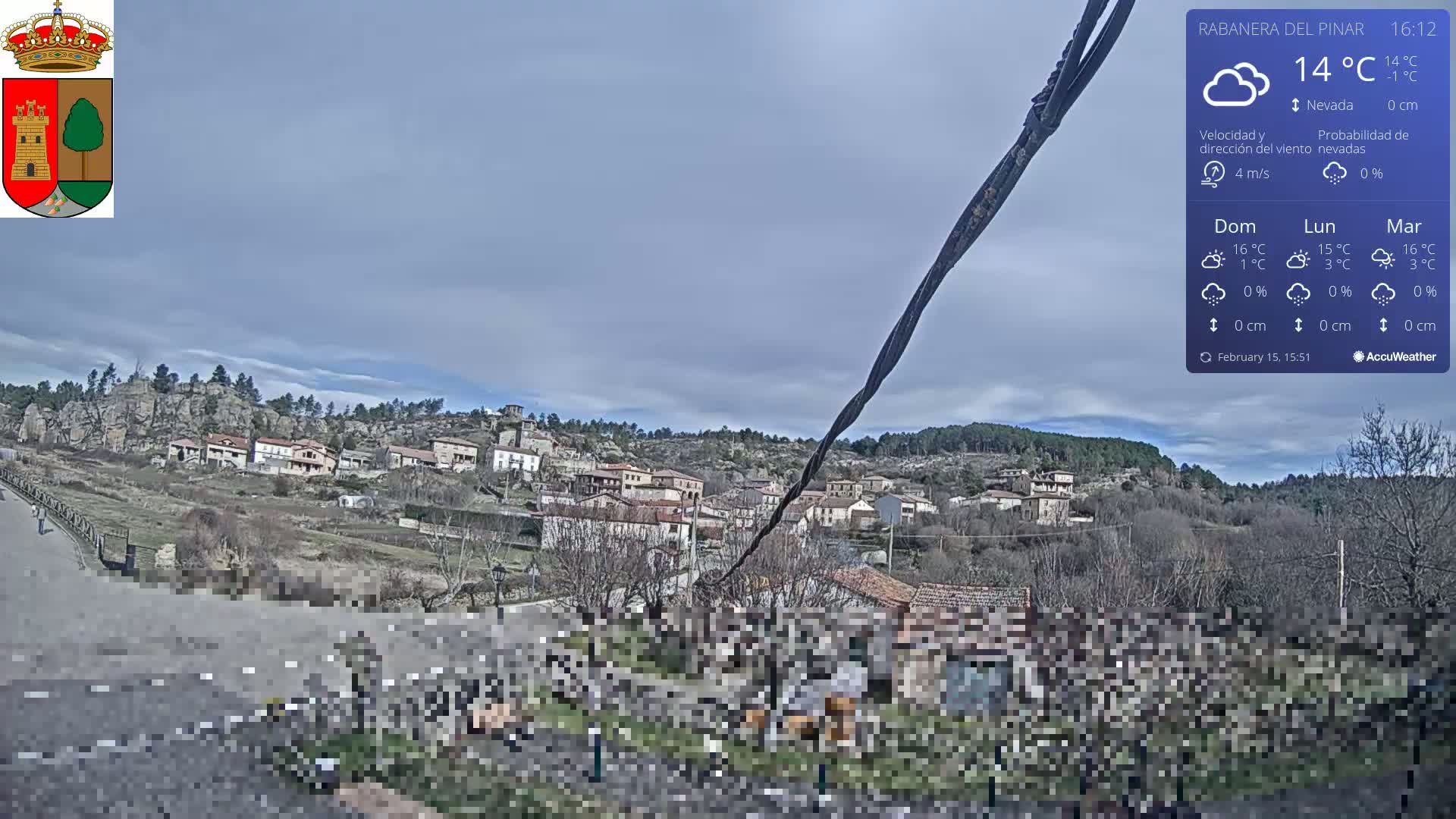 A mostly cloudy day reveals a small village nestled in a hillside, with leafless trees and a road in the foreground.