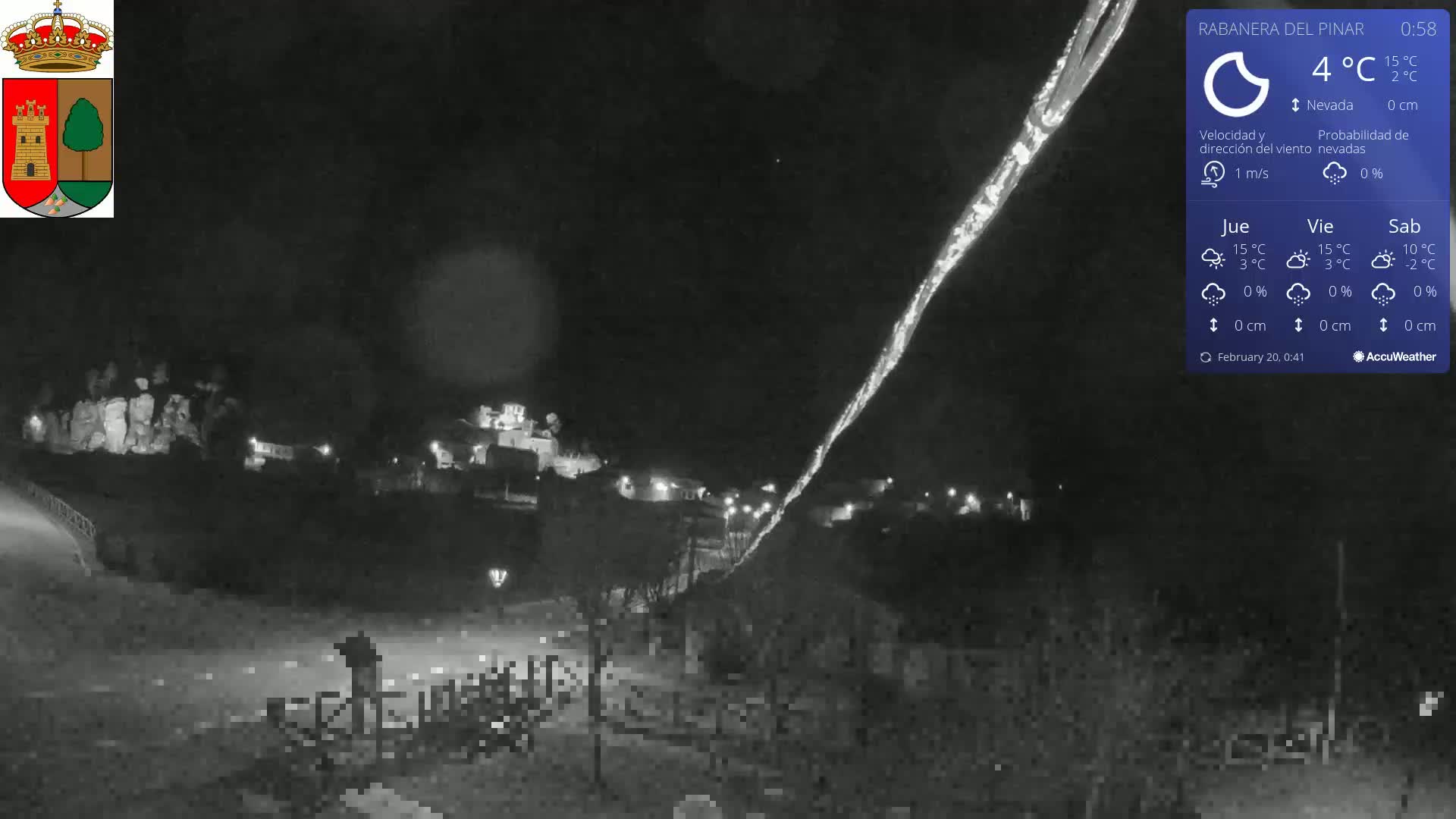 A nighttime view of a snow-covered town illuminated by scattered lights, under a dark sky with light snowfall.