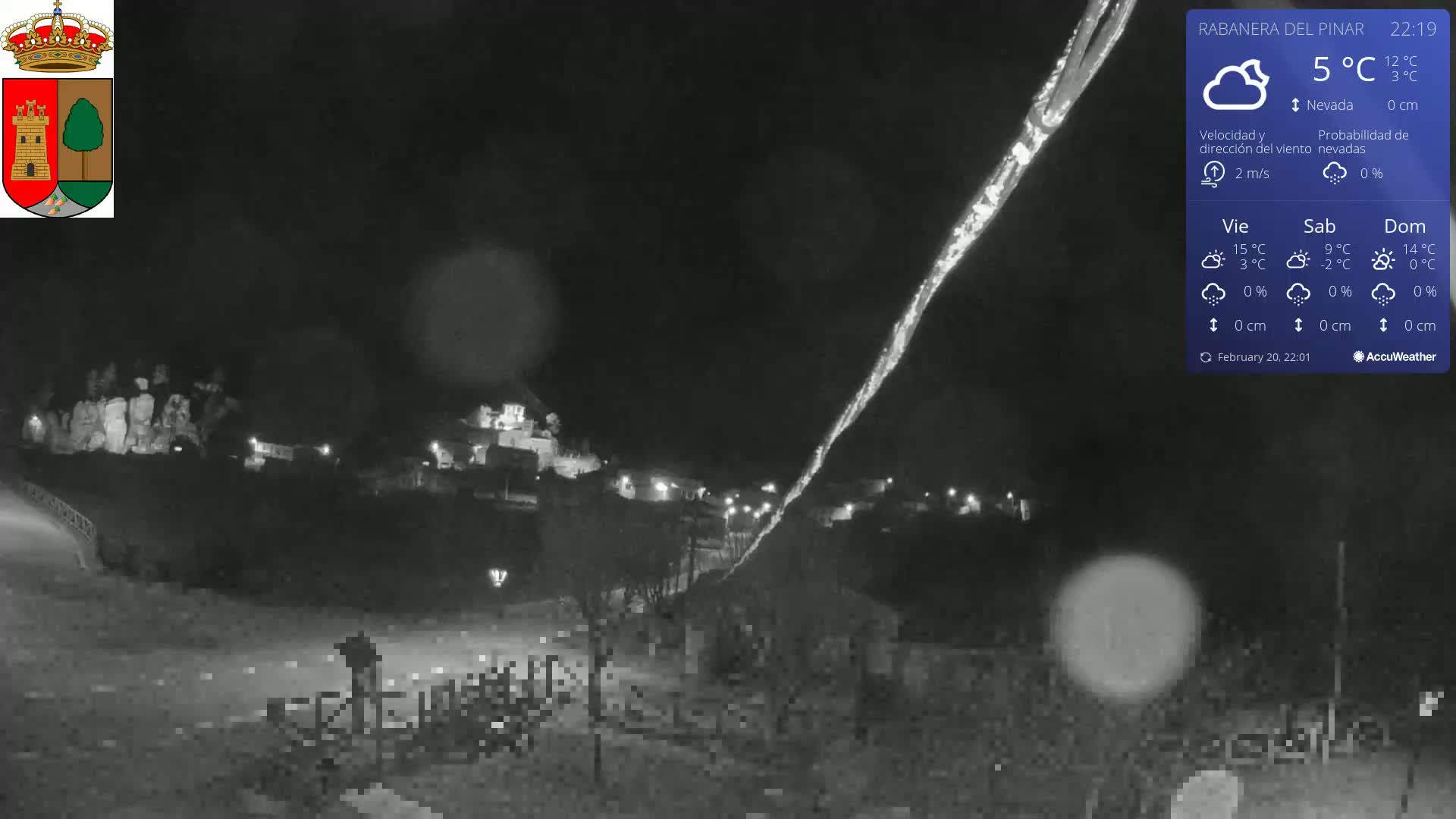 A nighttime, snowy scene shows a small town illuminated by lights, with a bright streak possibly from car headlights cutting across the dark sky.