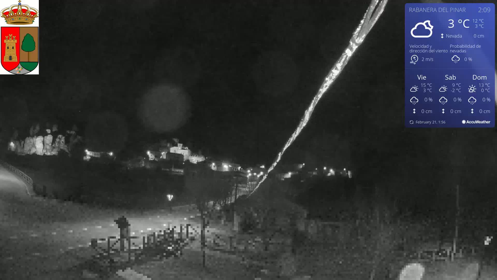 A nighttime, snowy scene shows a small village illuminated with faint lights, under a dark sky with a bright streak possibly caused by a light source.