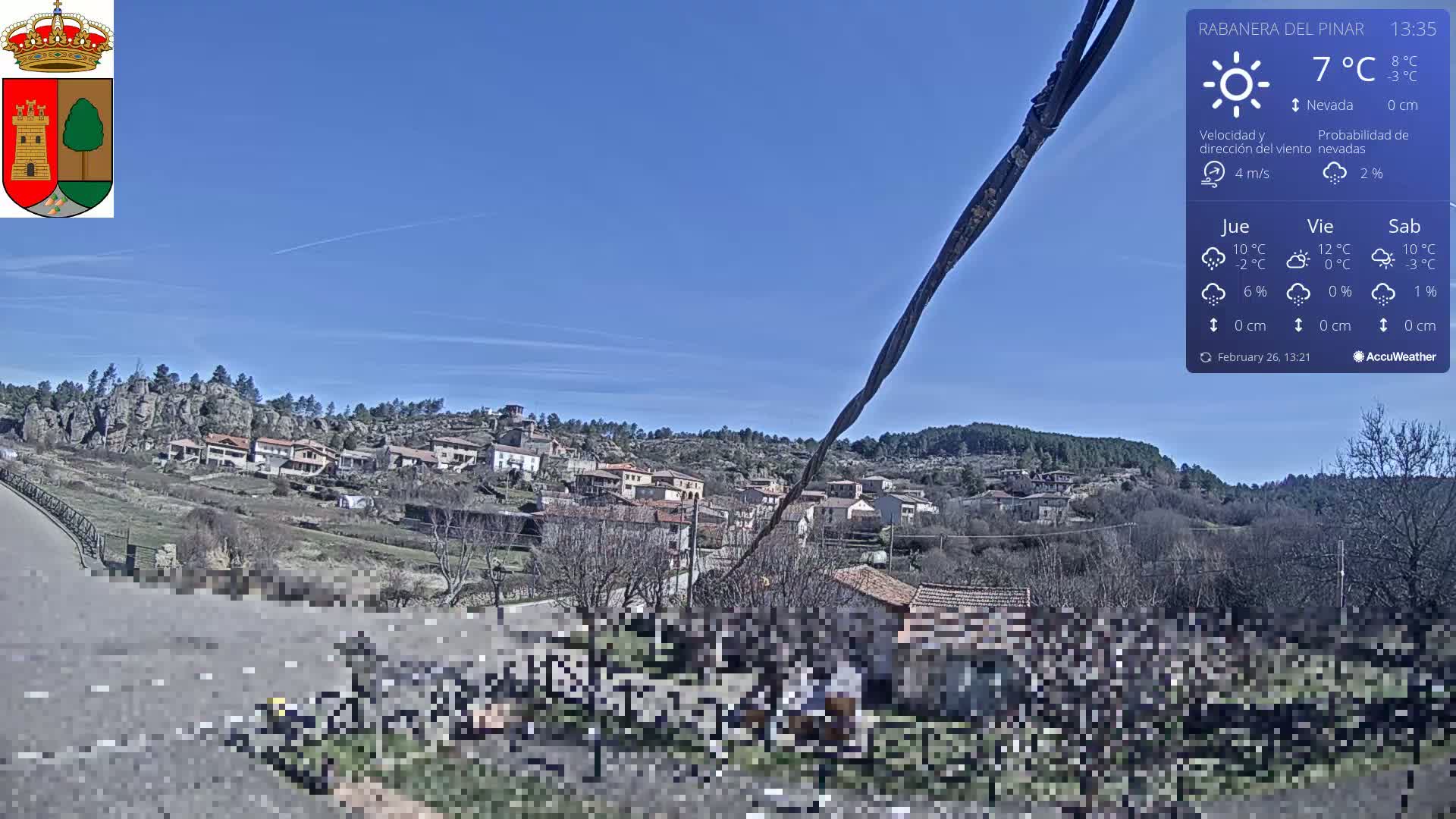 A small village nestled in a hilly landscape under a clear blue sky is viewed from a roadside, with a temperature of 7°C.