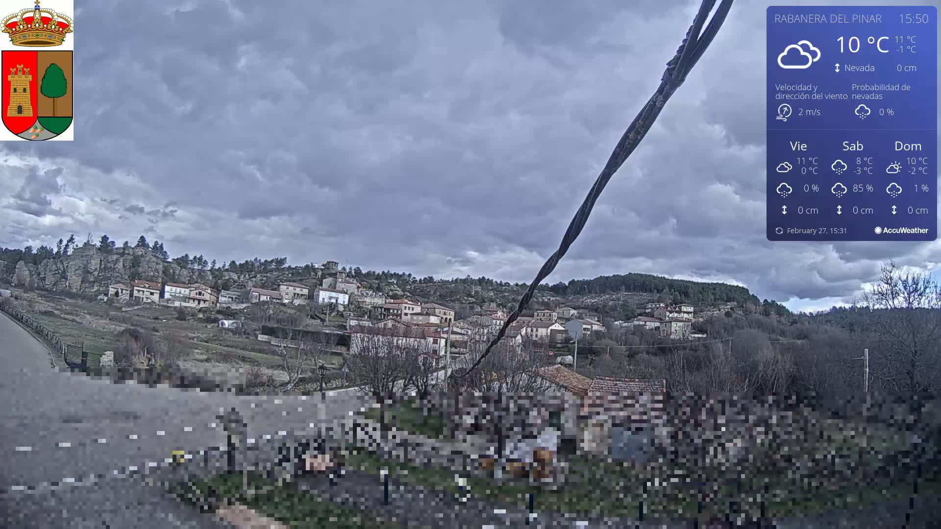 A small village nestled in a hilly landscape under a mostly cloudy sky is viewed from a slightly elevated position.