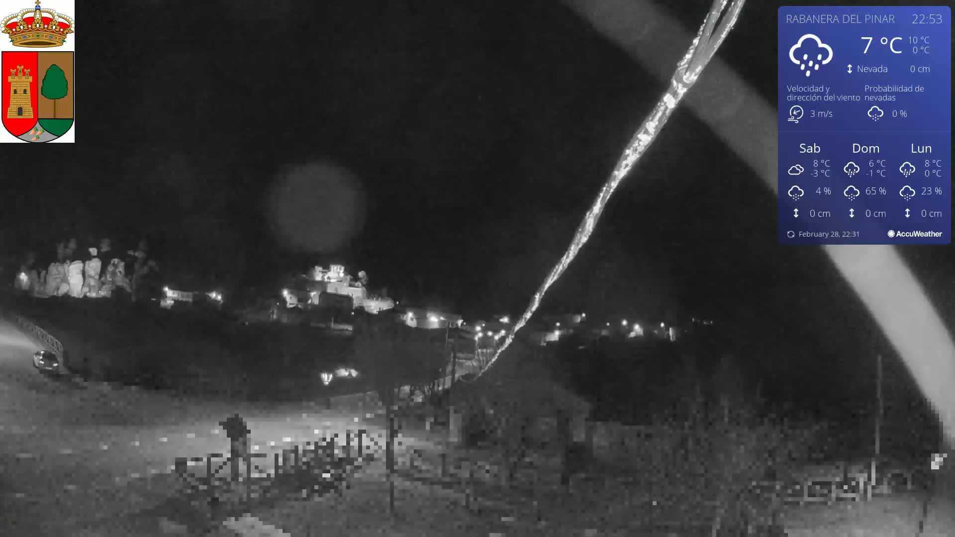 A nighttime, snowy scene shows a small town illuminated by scattered lights, with a person standing near a fence in the foreground.