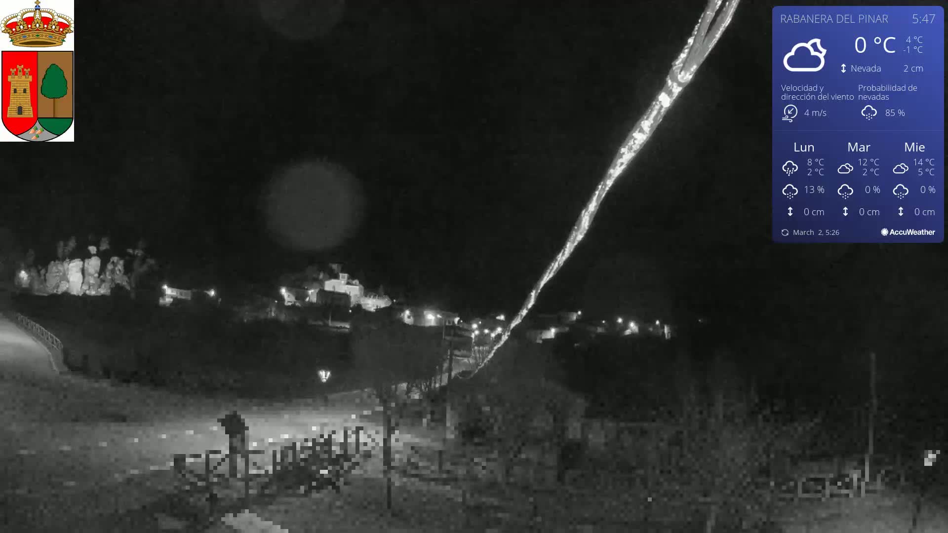 A nighttime, snowy scene shows a village illuminated by scattered lights, with a long, bright streak in the sky above.