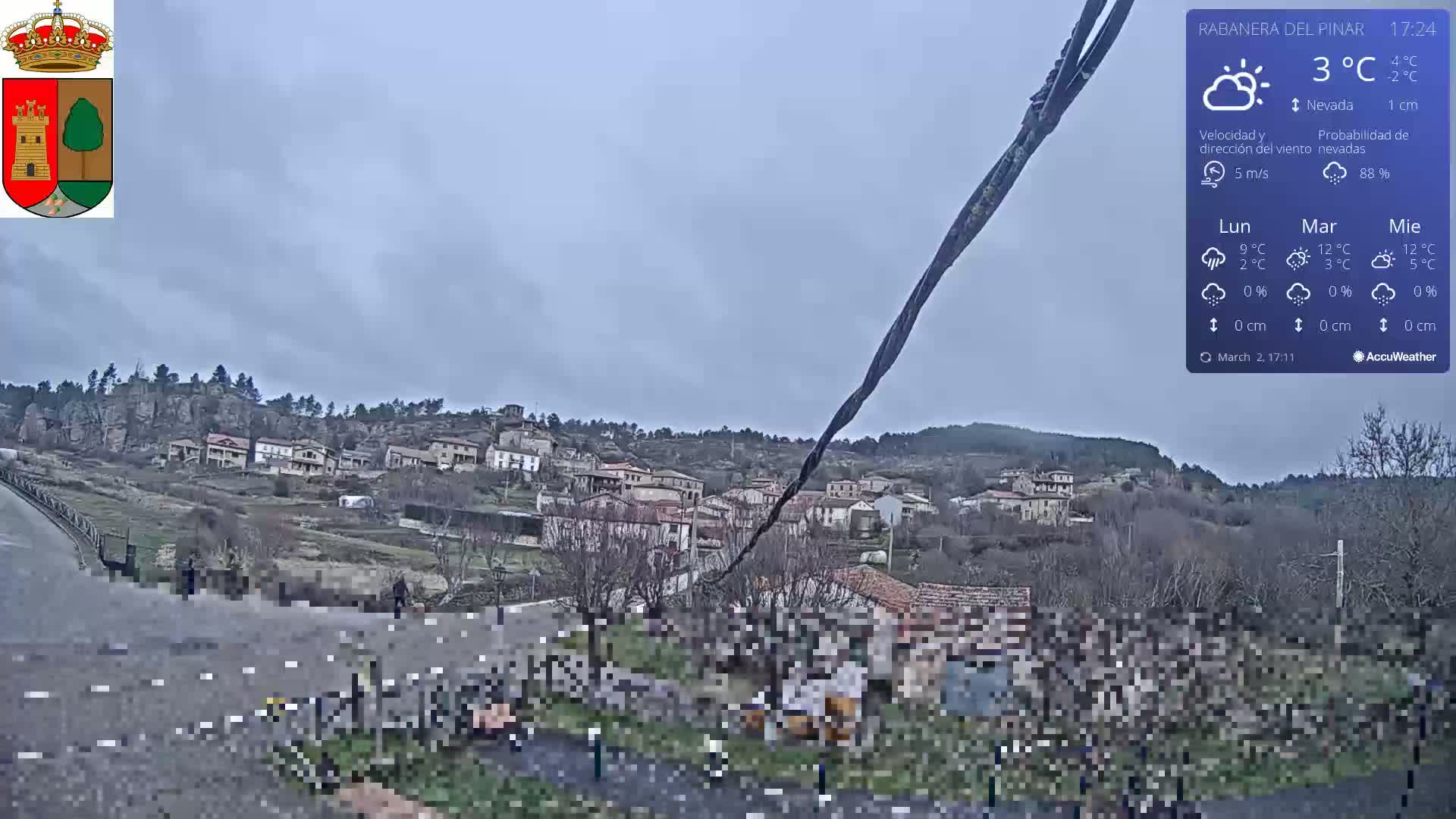 A small village nestled on a hillside under an overcast sky is viewed from a roadside, with a partly obscured road and some people gathered in the foreground.