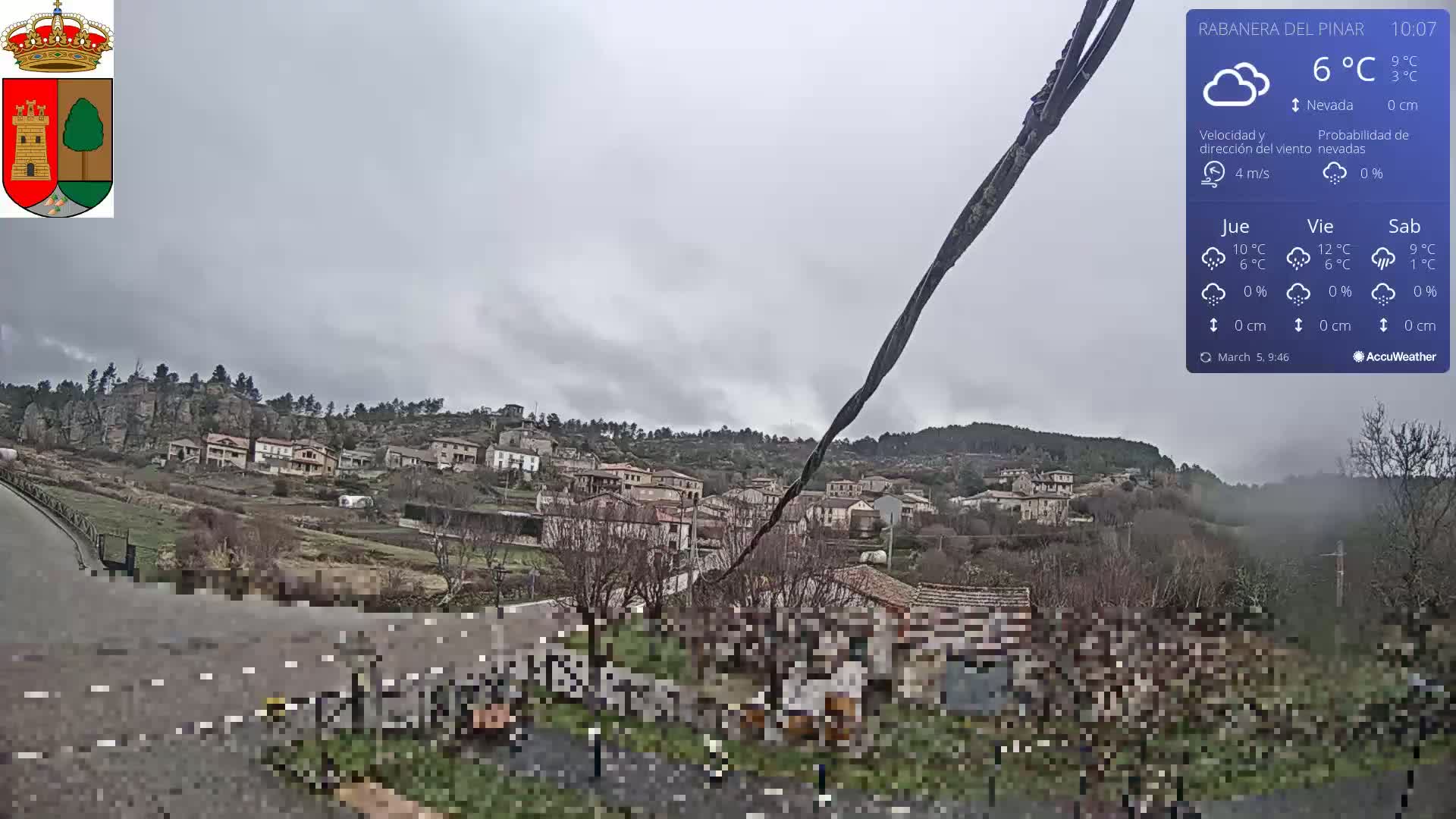 A small village nestled in a hillside is viewed from a slightly elevated position on an overcast day with a 6°C temperature.
