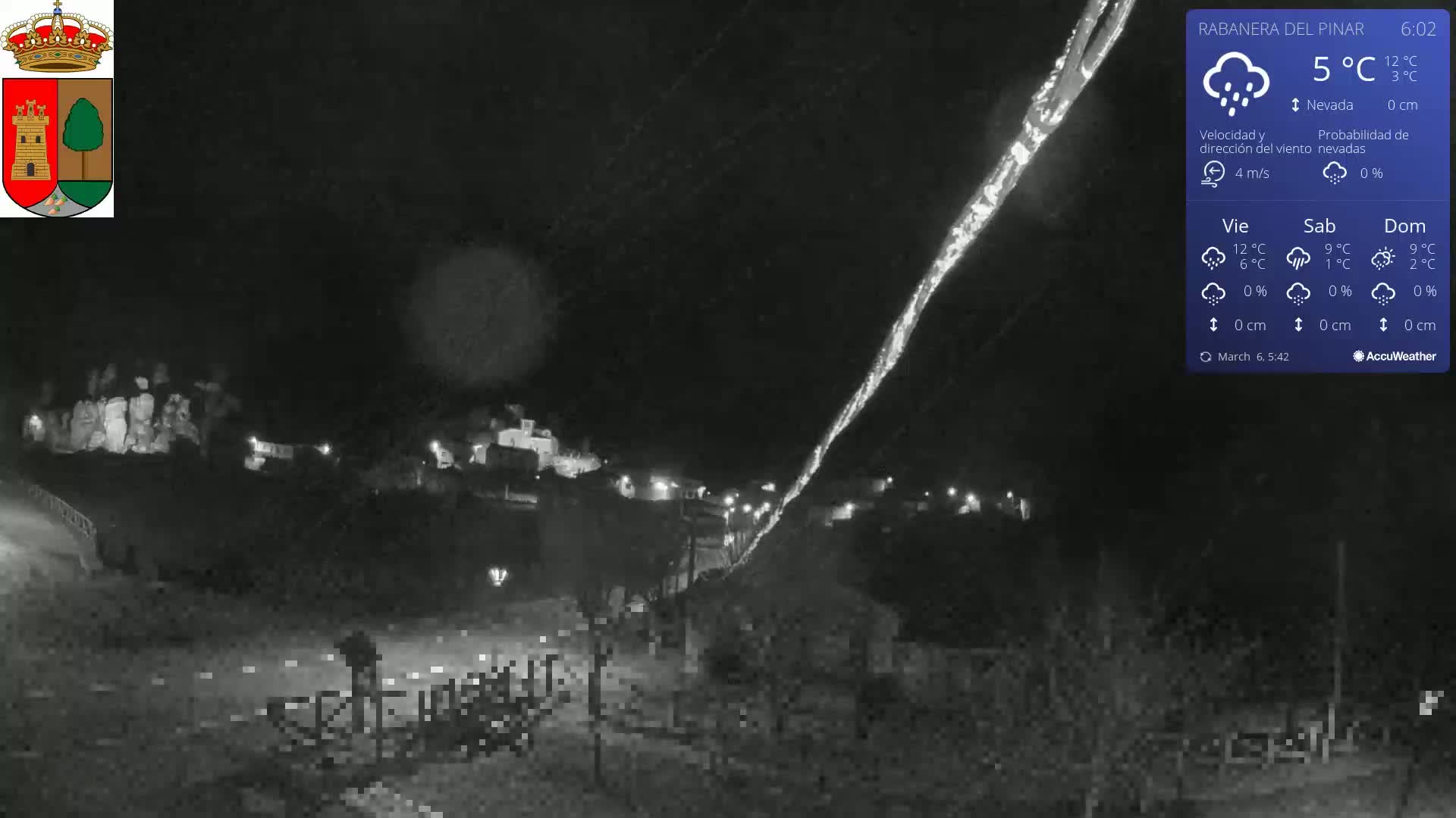 A nighttime, snowy scene shows a small town nestled in a valley, illuminated by scattered lights, under a dark sky with a faint light trail.