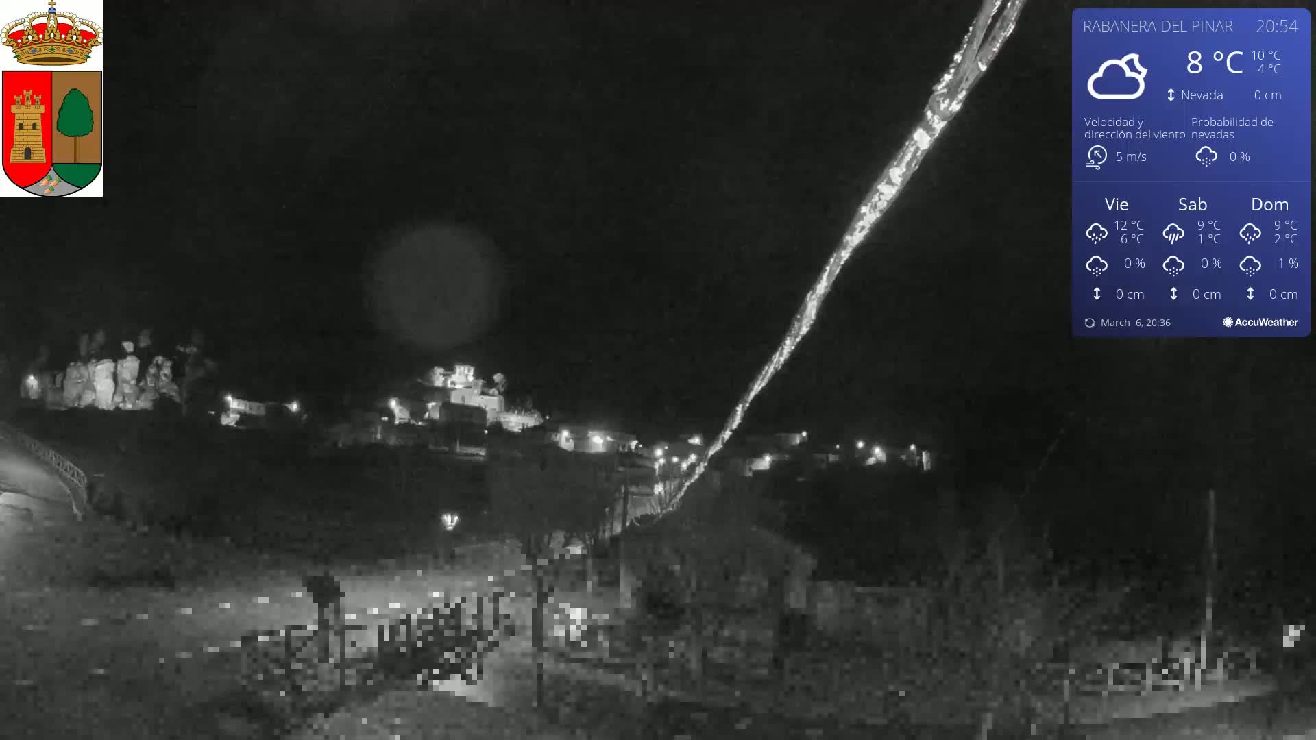 A nighttime, snowy scene shows a small village illuminated by scattered lights, with a long, bright streak descending from the upper right corner of the image.