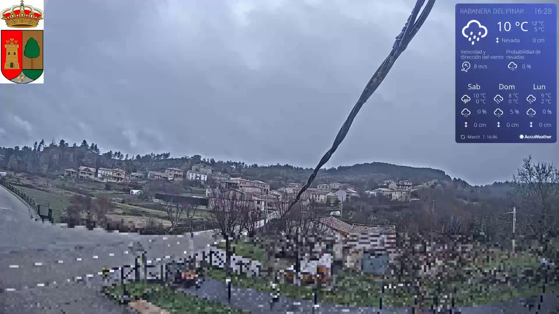 A small village nestled on a hillside is viewed from a slightly elevated position on an overcast day with light rain.