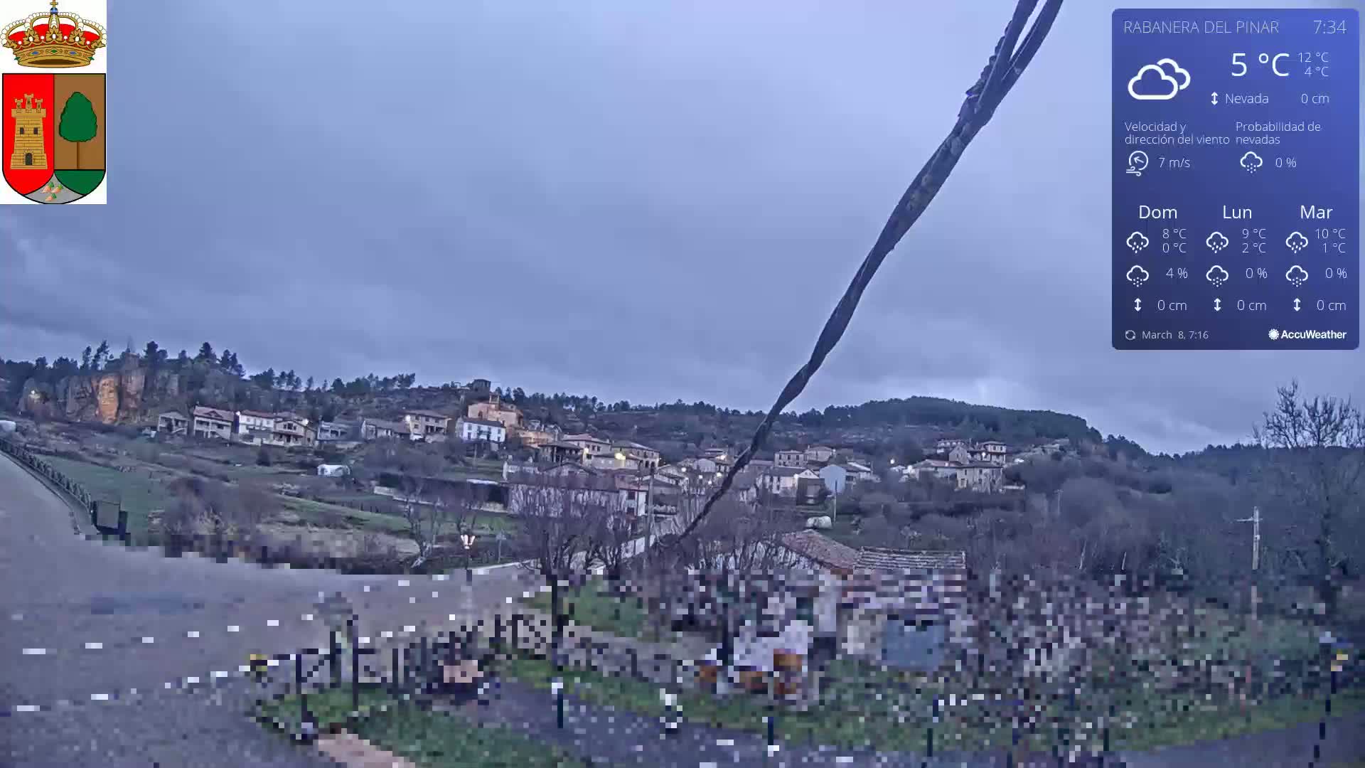 A small village nestled on a hillside under a cloudy, overcast sky is seen from a roadside vantage point.