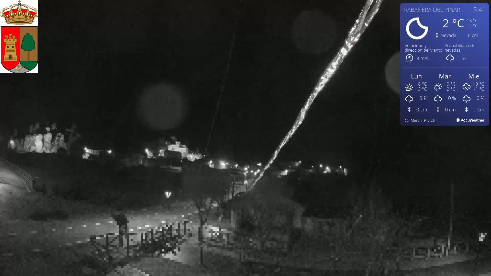 A nighttime view of a small town illuminated by scattered lights, under a dark sky with a light snow falling.