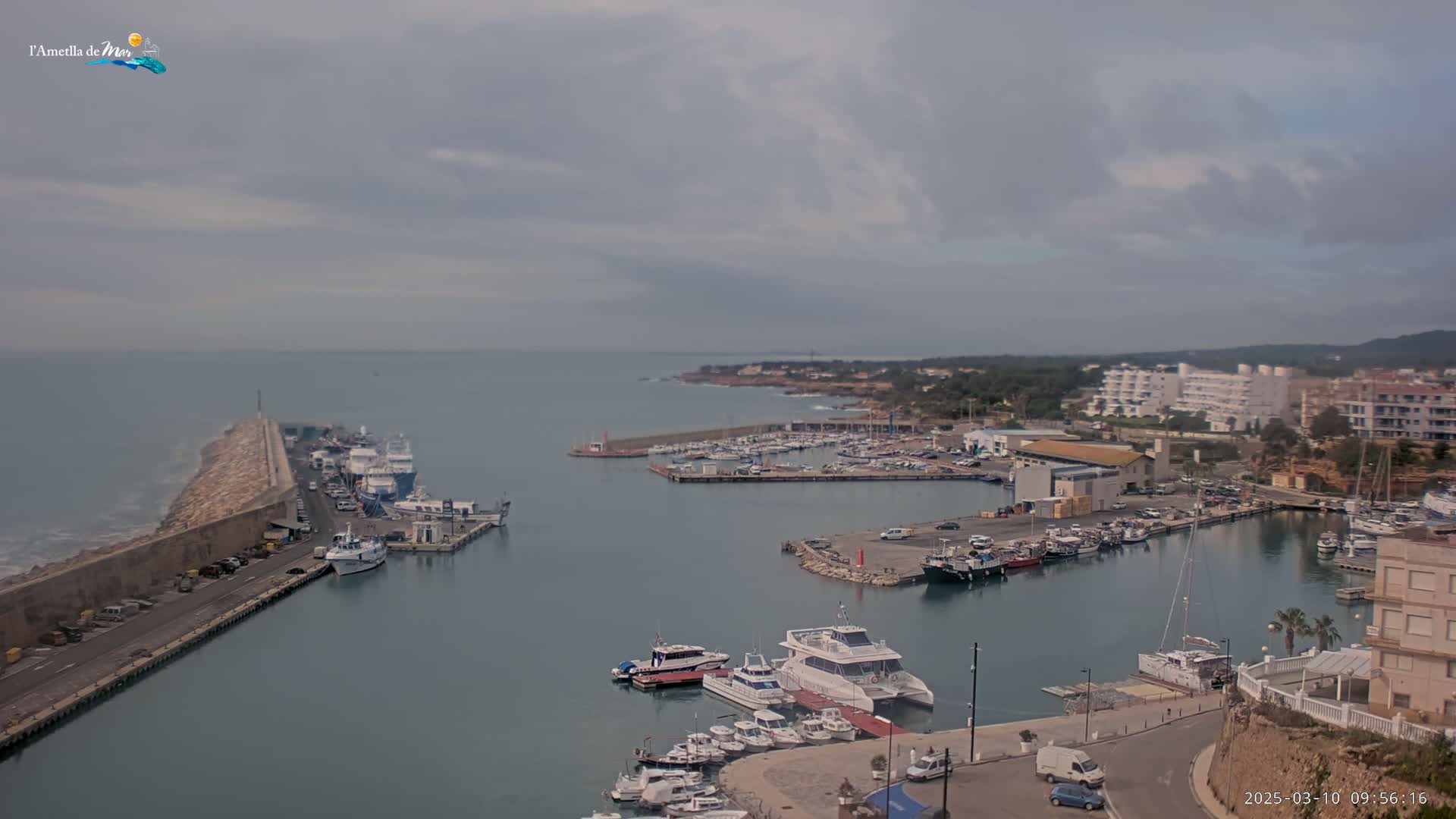 A harbor filled with boats is seen under a cloudy sky, with a long stone wall extending into the sea.