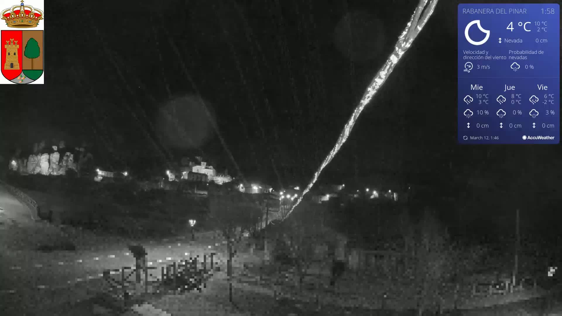 A nighttime, snowy scene shows a small town illuminated with faint lights, under a dark sky with falling snow.