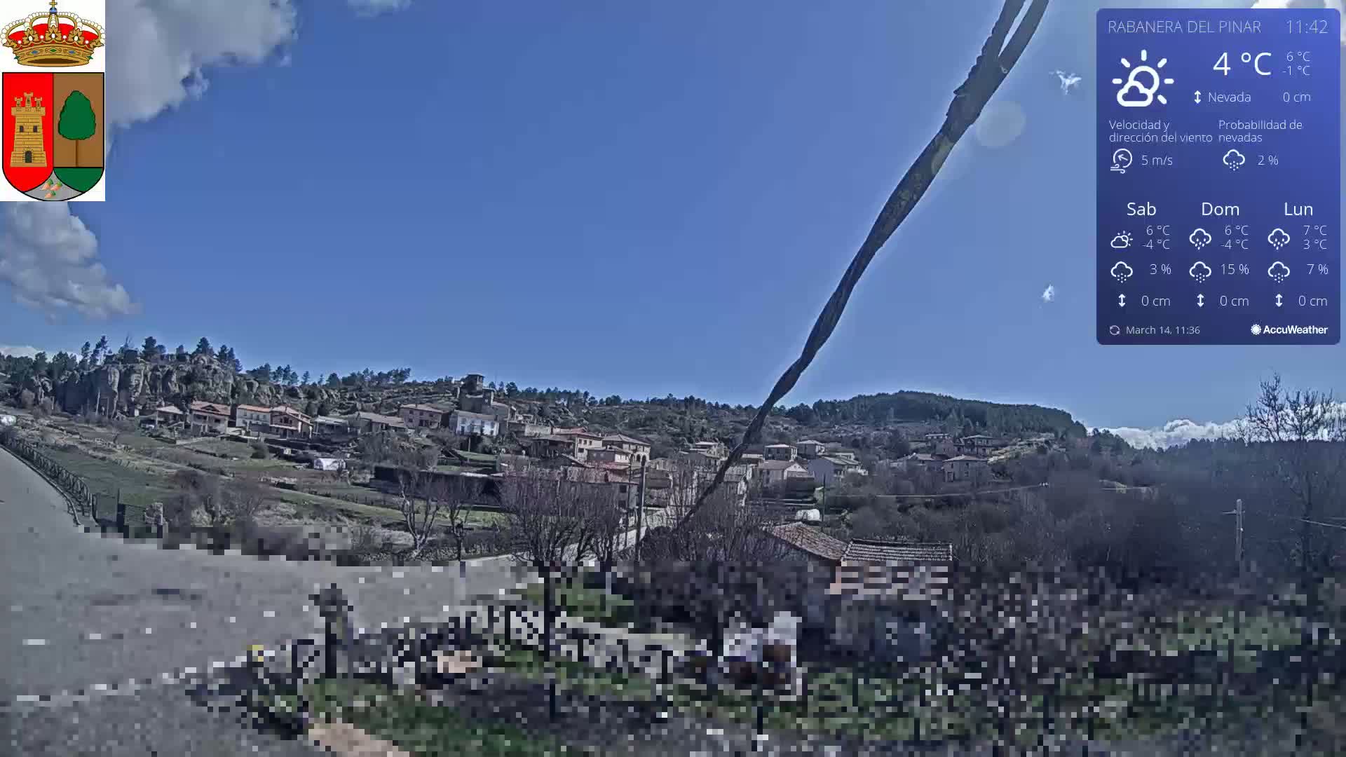 A mostly sunny day reveals a small village nestled in a hillside, with sparse trees and some snow-covered areas visible in the distance.