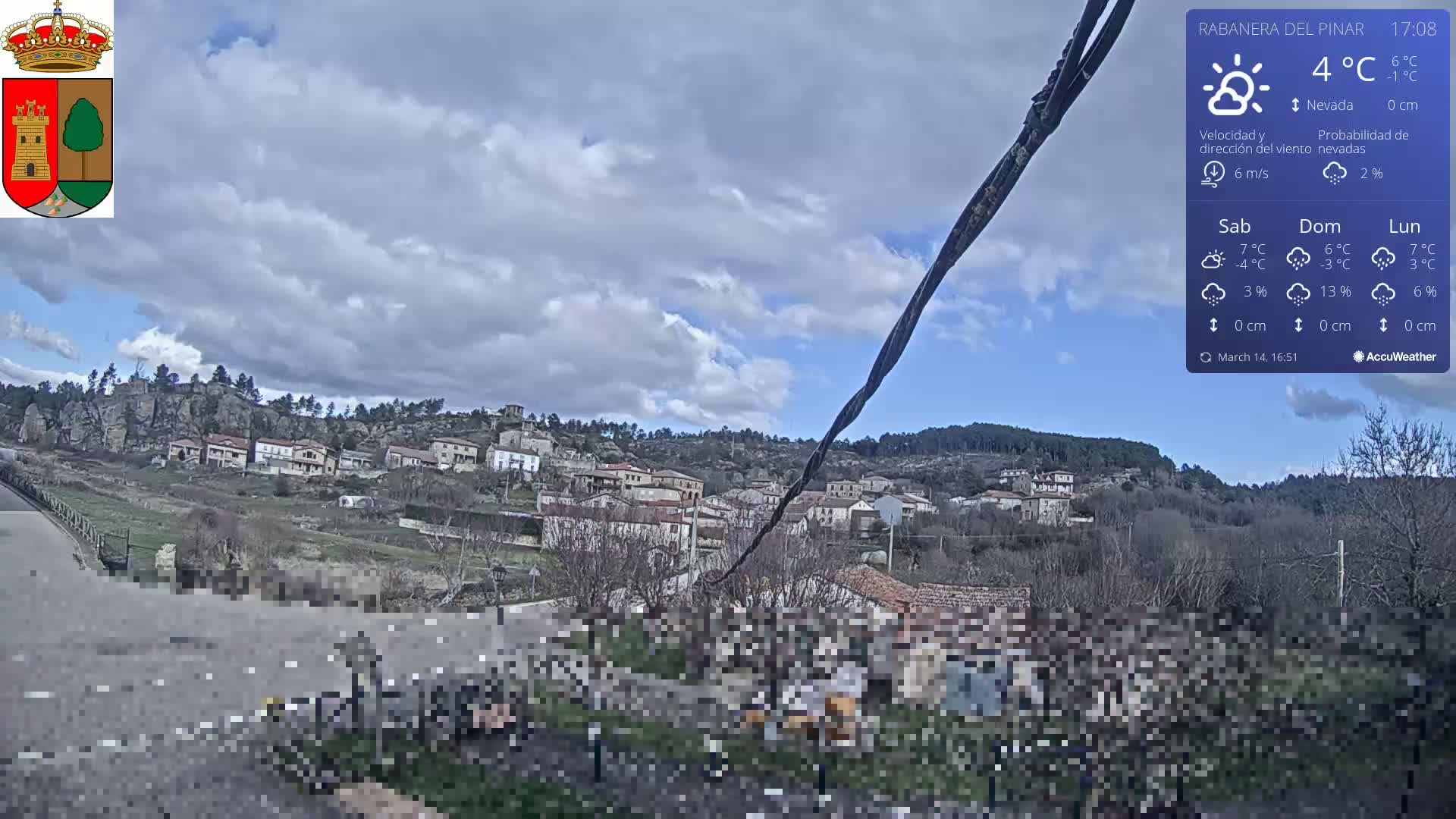 A partly cloudy sky over a small village nestled on a hillside, with a road and some people visible in the foreground.