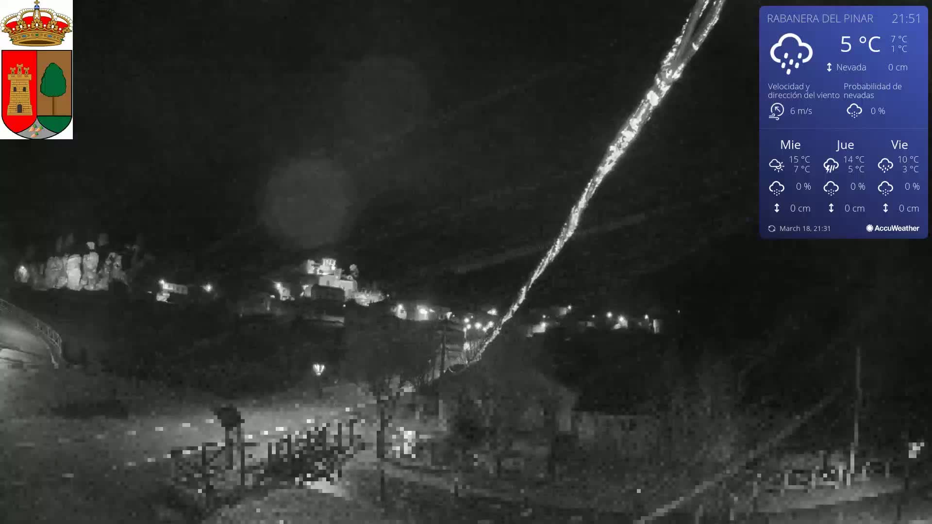 A nighttime view of a small town illuminated by scattered lights, with light rain or snow falling under a dark sky.