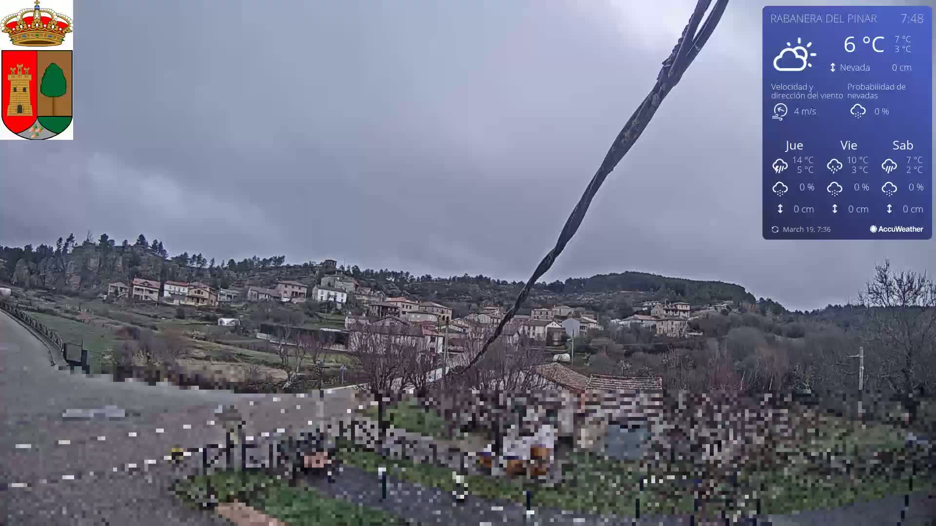 A small village nestled in a hilly landscape under an overcast sky is viewed from a roadside vantage point.