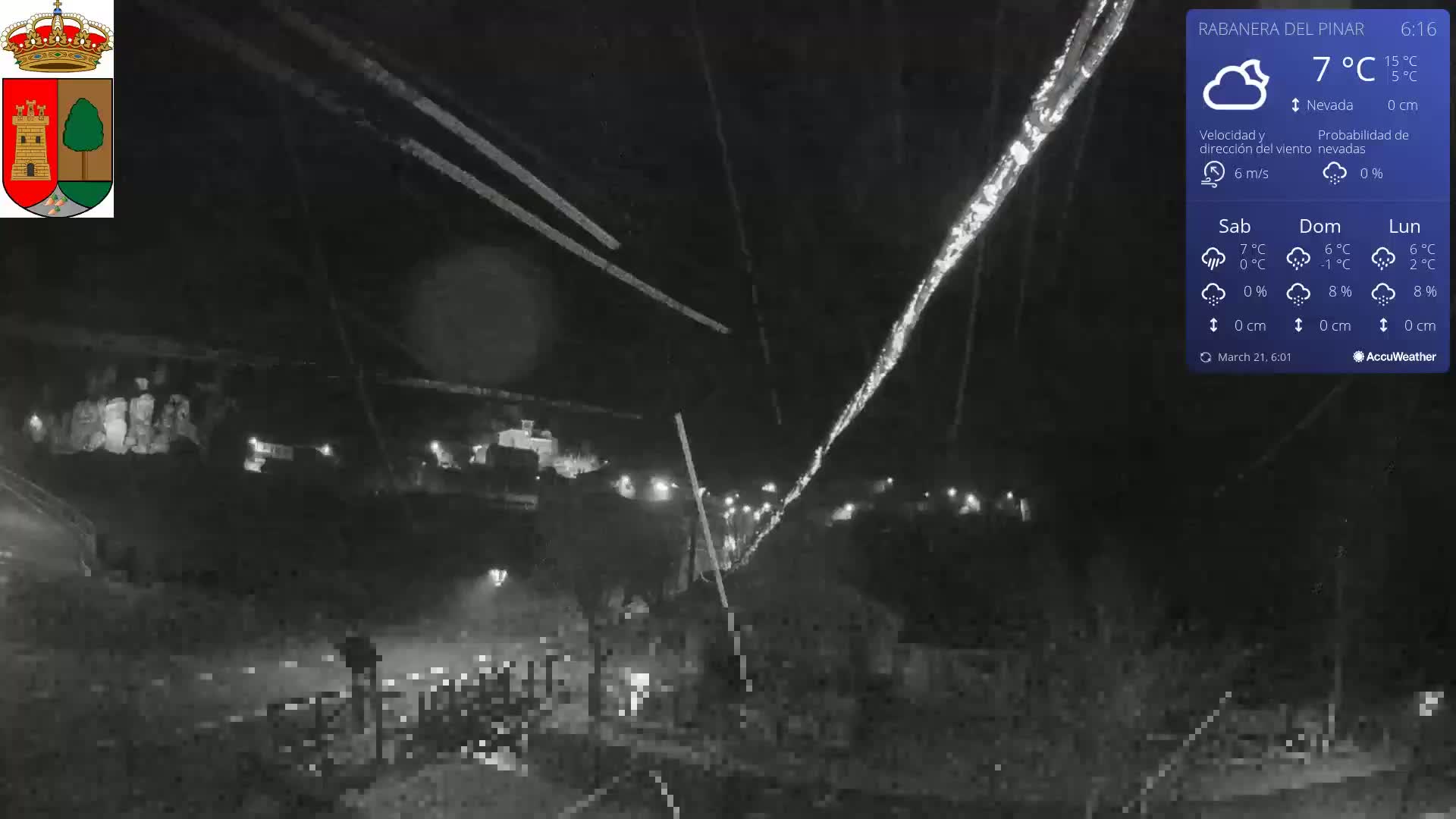 A nighttime, low-light view of a snow-covered area with some lights visible in the distance, under a cloudy sky with a light wind.