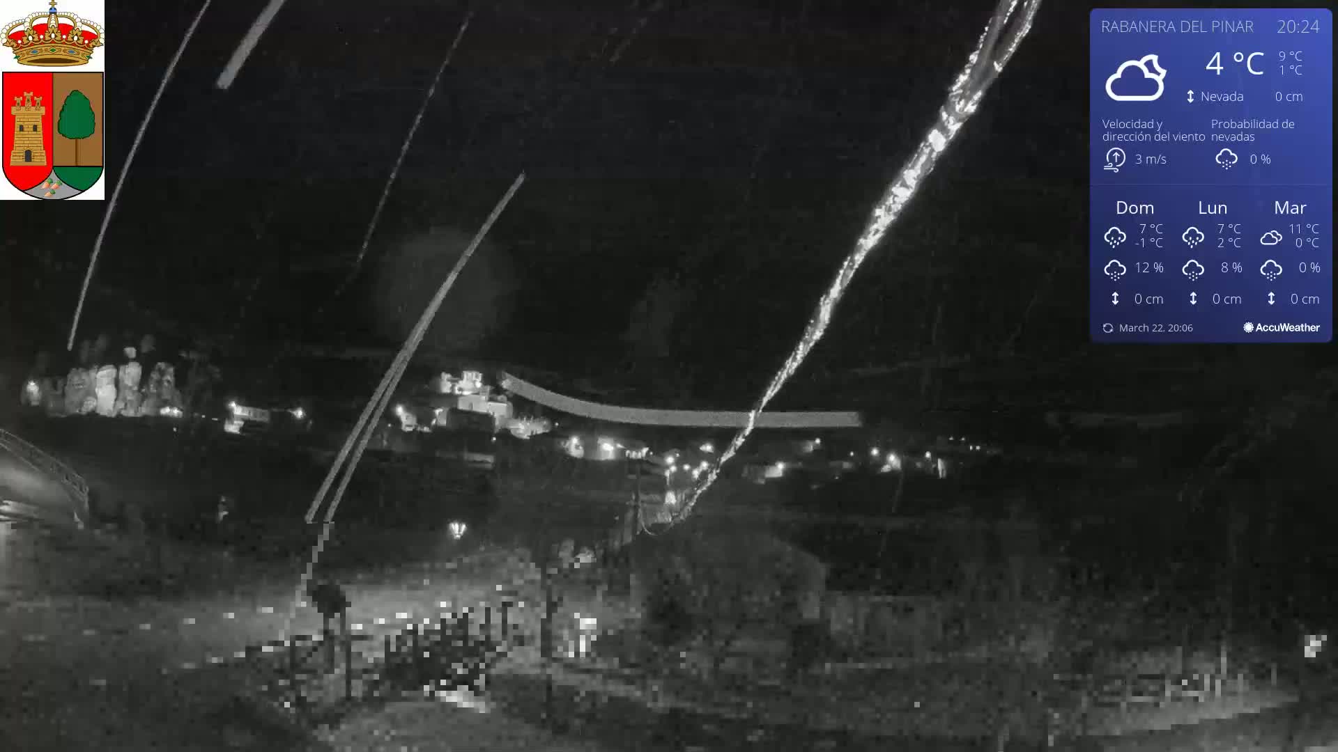 A nighttime, low-light view of a snow-covered area with some lights visible in the distance, under snowy conditions.