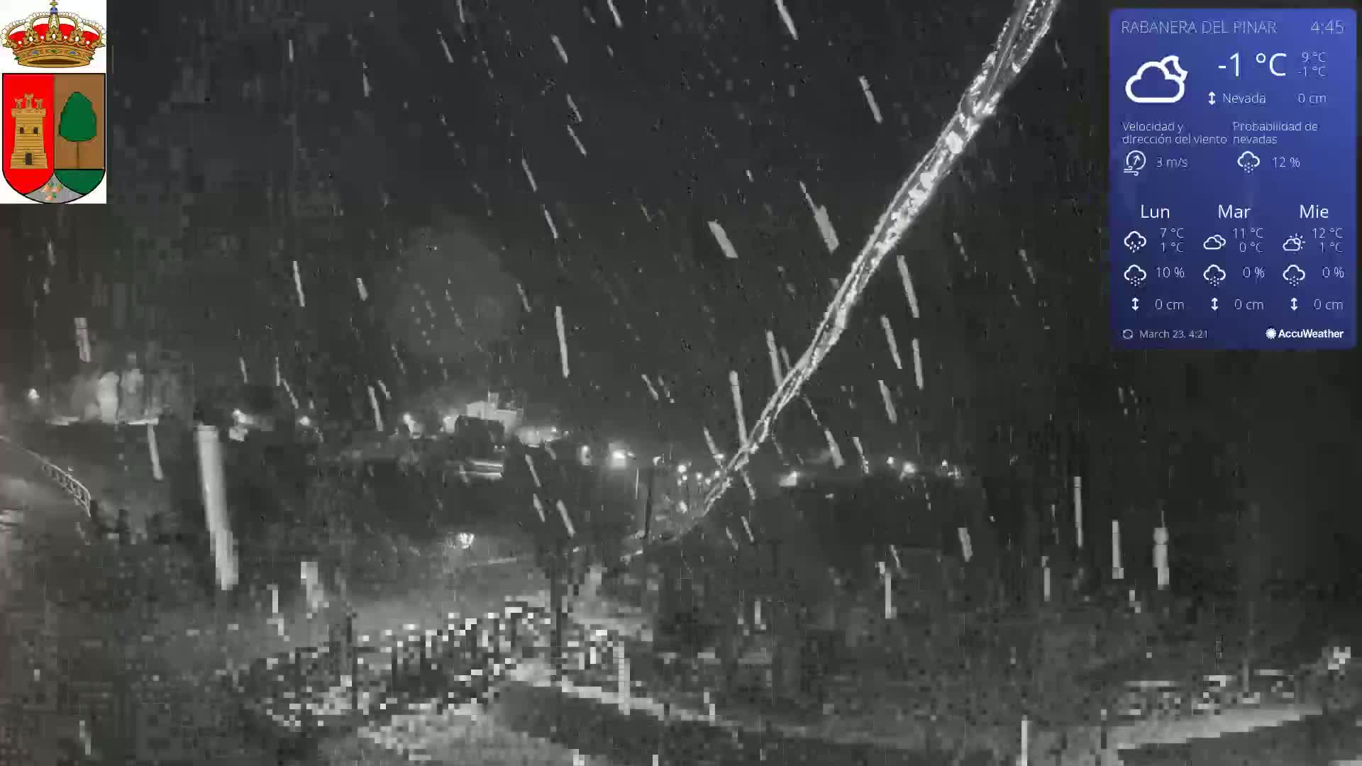 A nighttime scene of snow falling over a dark, sparsely lit area with some buildings visible in the distance.