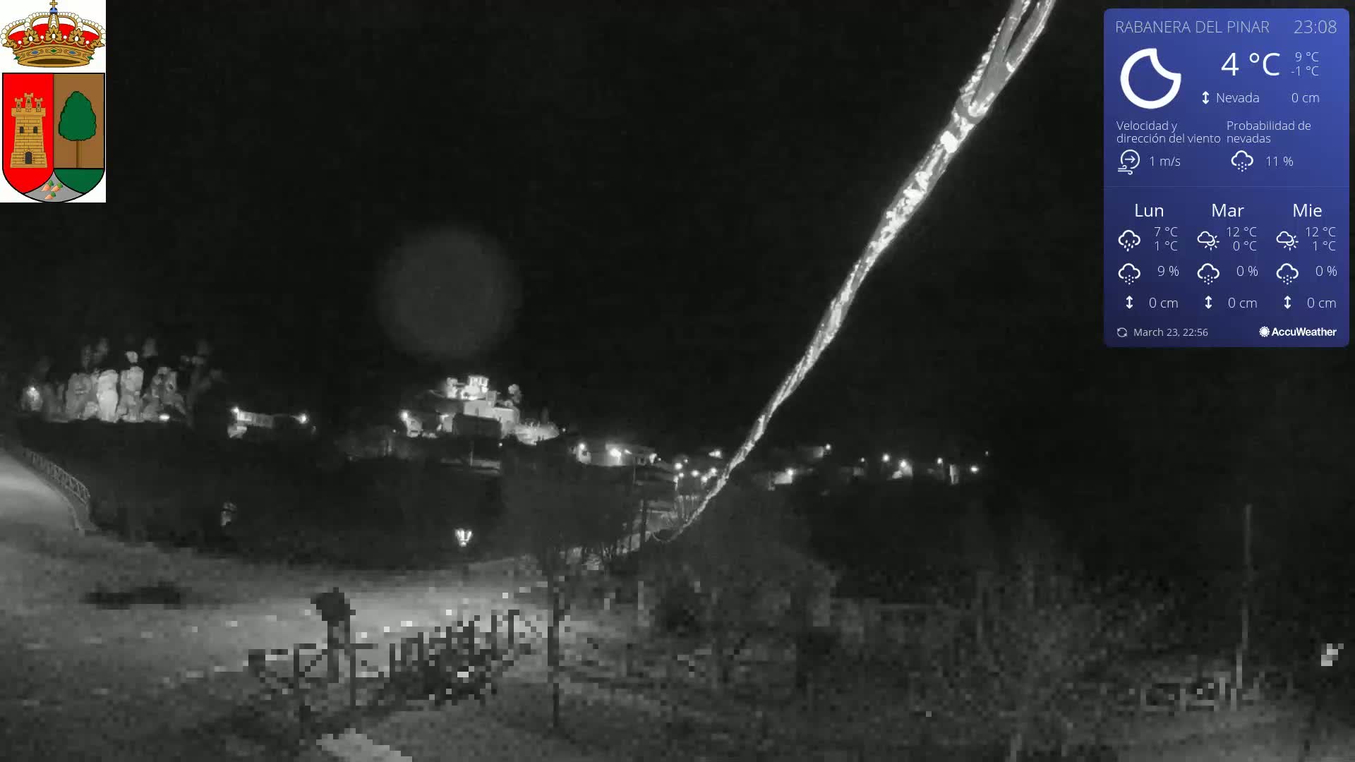 A nighttime, snow-covered landscape shows a small village with faint lights under a dark sky, with a thin streak of light possibly from a meteor or searchlight.