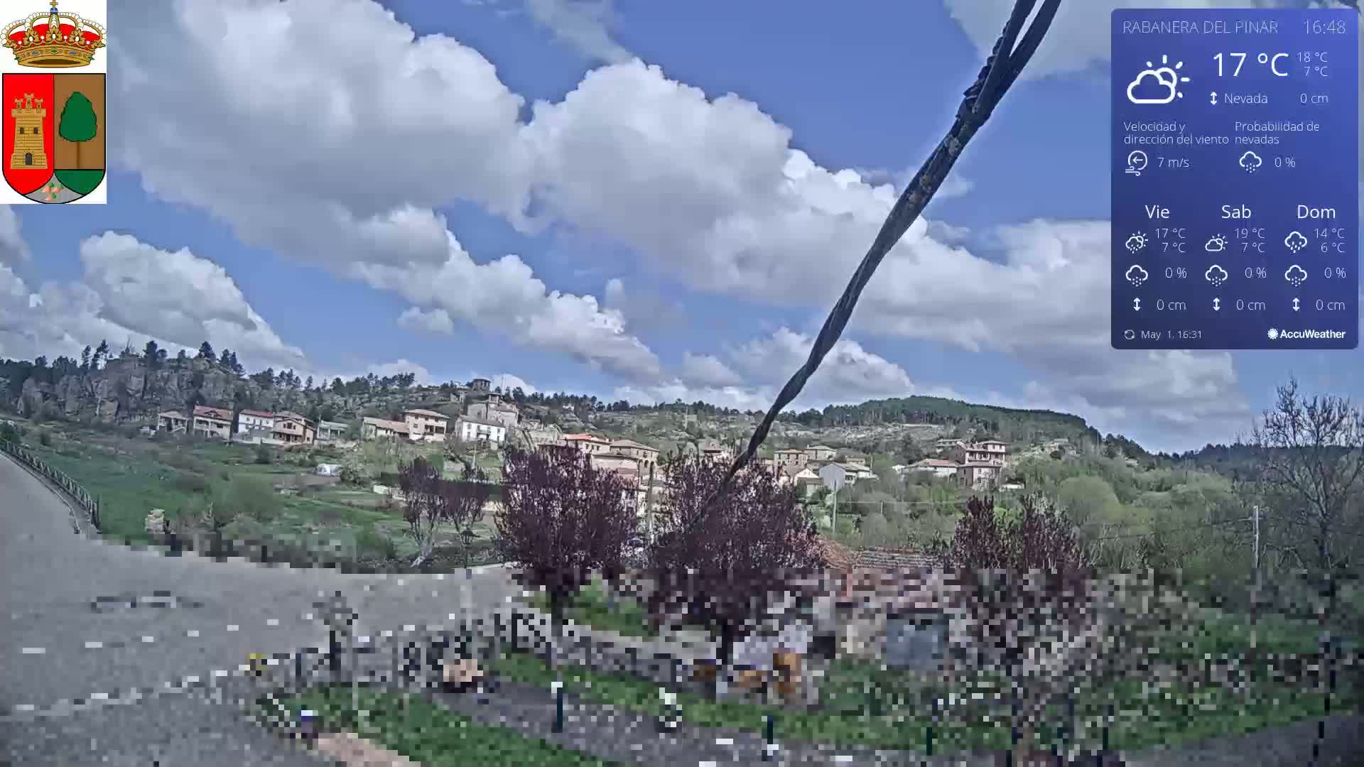 A partly cloudy sky over a small village nestled in a hillside, with people gathered near a road in the foreground.
