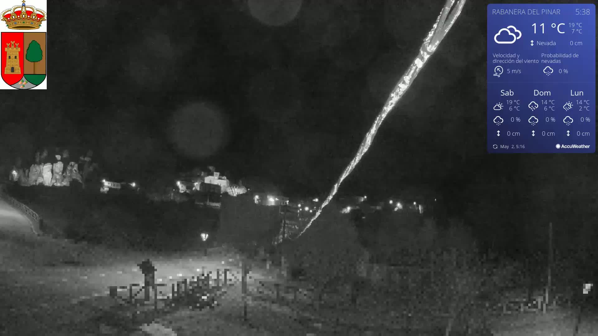 A nighttime, snowy scene shows a village in the distance, illuminated by faint lights, with a long, bright streak in the sky.