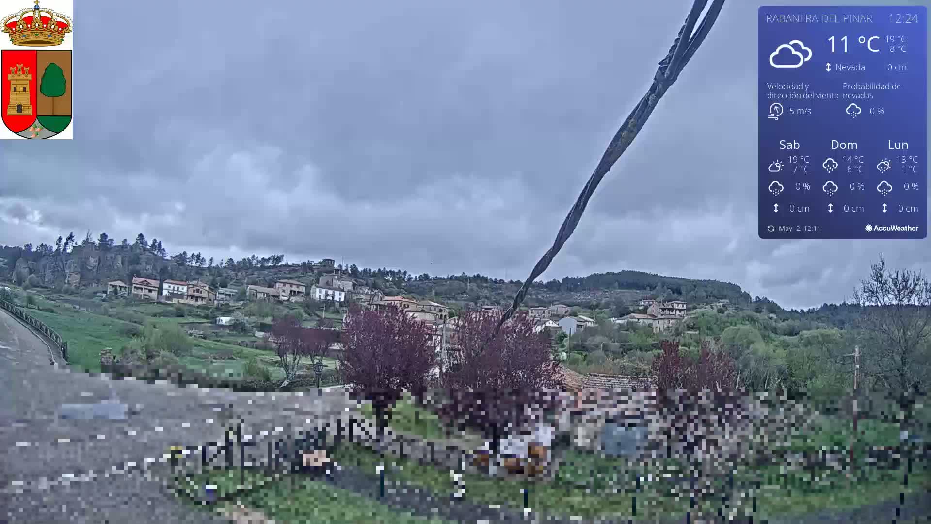 A small village nestled in a hilly landscape under an overcast sky is viewed from a roadside vantage point.