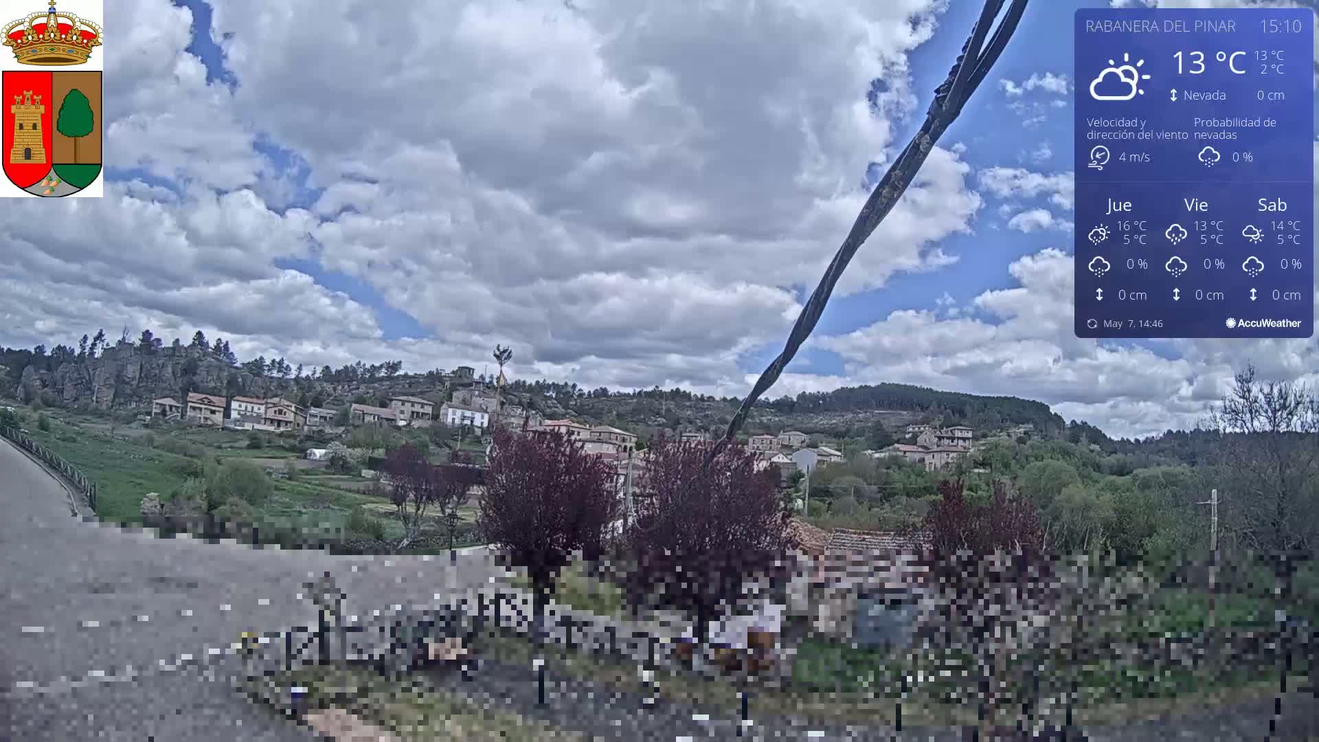 A partly cloudy sky hangs over a small village nestled on a hillside, with a few dark-colored trees in the foreground.
