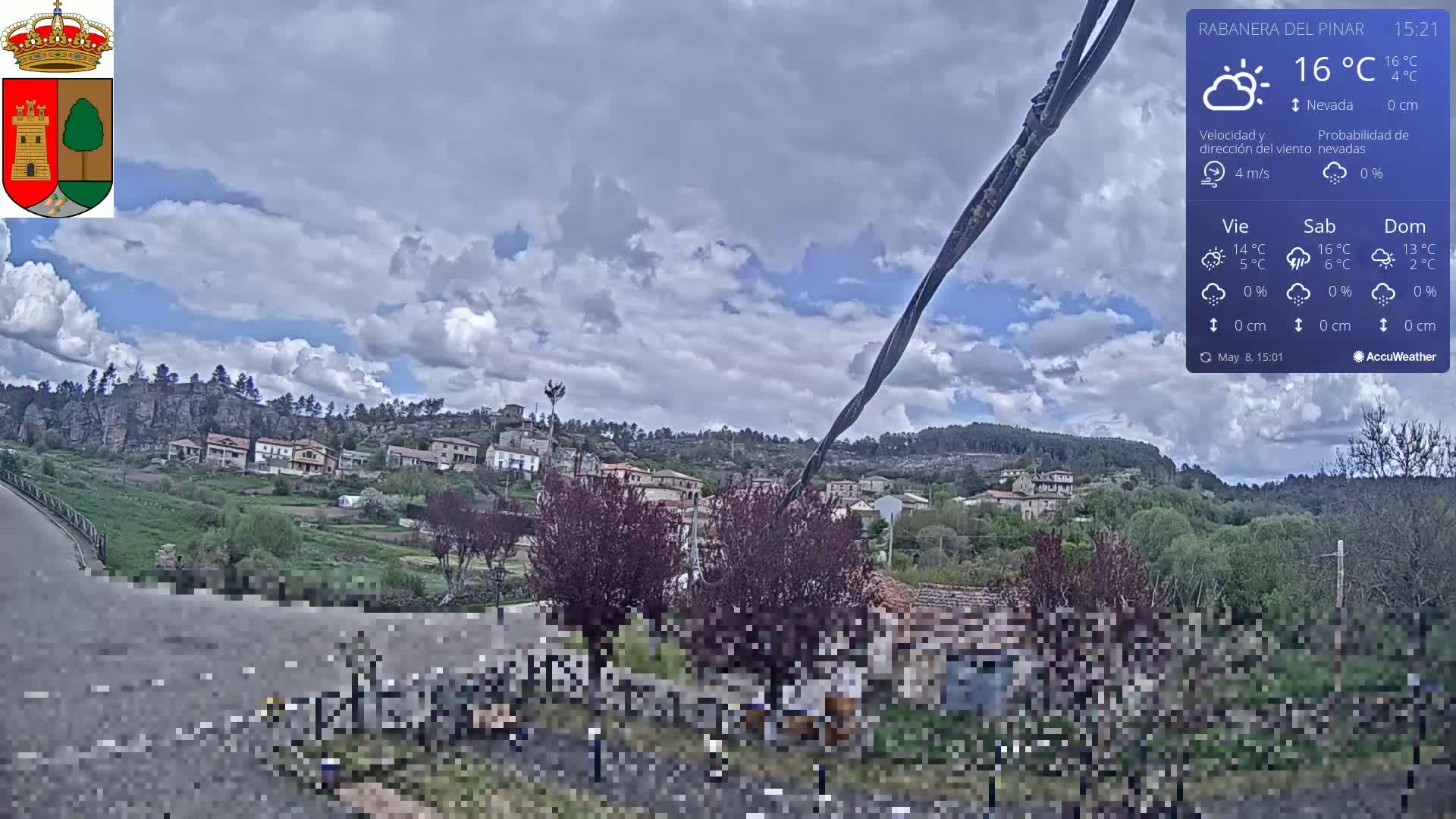 A partly cloudy sky hangs over a small village nestled on a hillside, with a road and some people visible in the foreground.