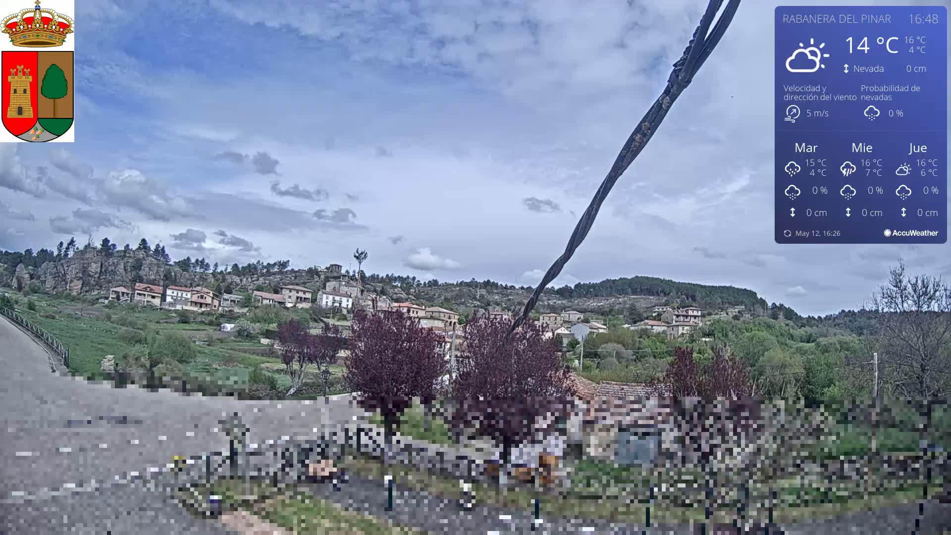 A partly cloudy sky hangs over a small village nestled on a hillside, with a road and some vegetation in the foreground.