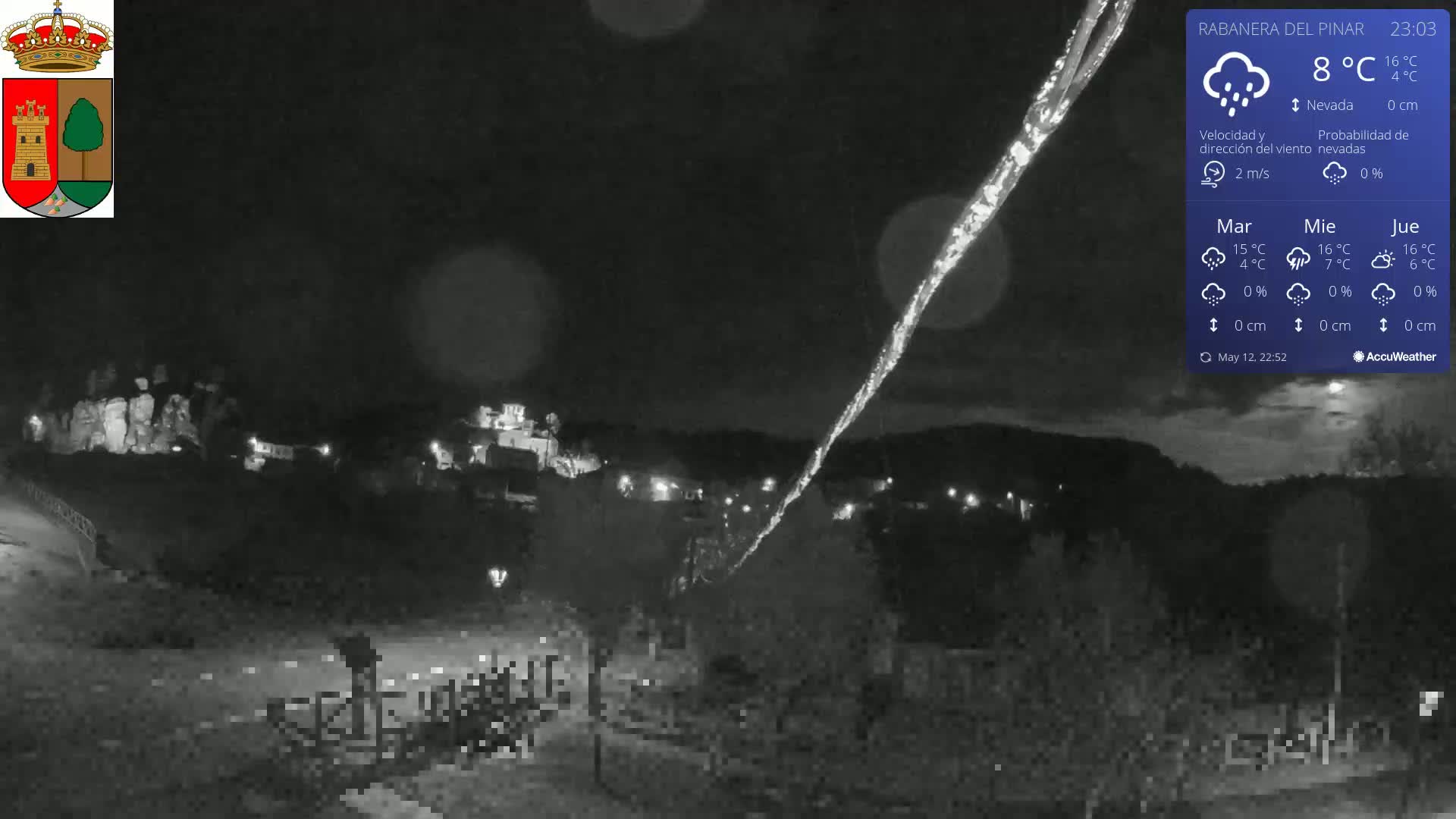 A nighttime, low-light view of a small town under a slightly overcast sky with light rain shows a long, bright streak in the sky.