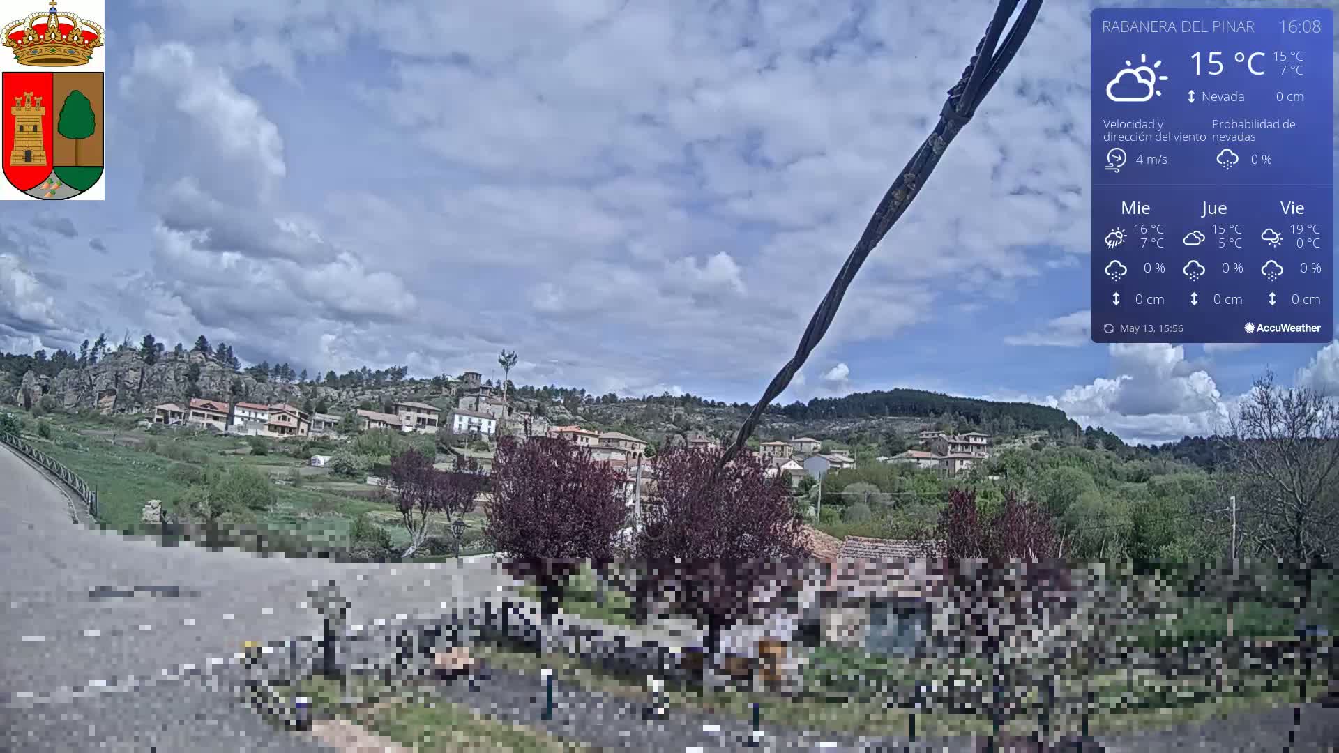 A partly cloudy sky hangs over a small village nestled in a hilly, green landscape.