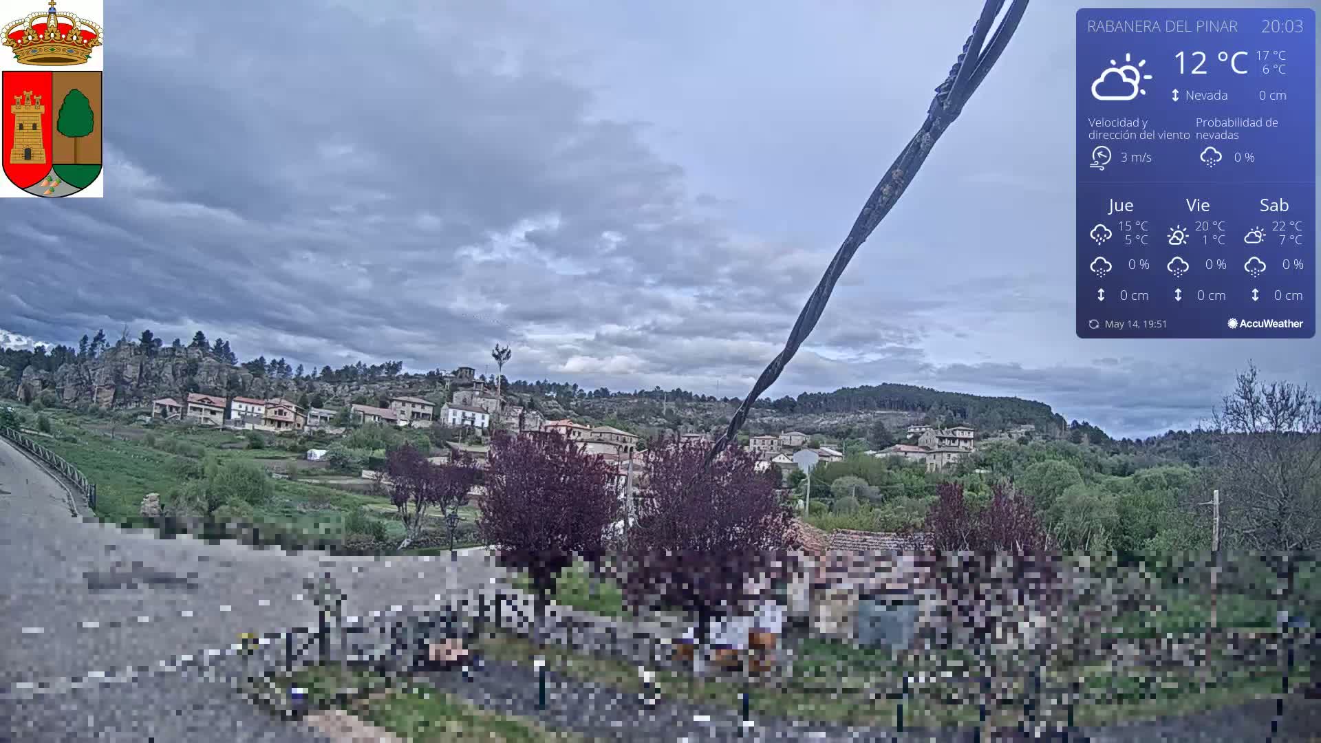 A small village nestled in a hilly landscape under a mostly cloudy sky is viewed from a slightly elevated vantage point.