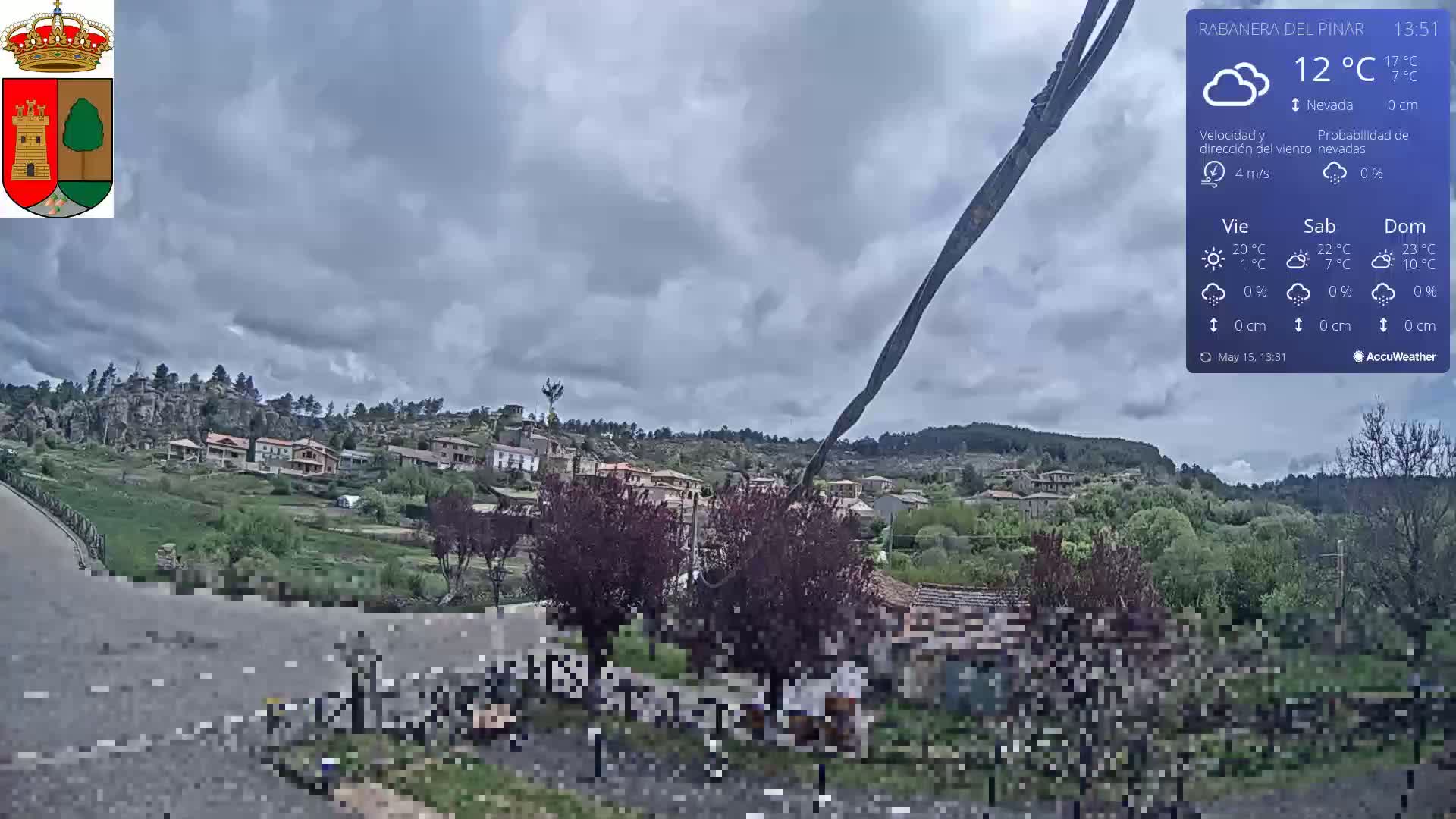 A small village nestled in a hilly landscape under a cloudy sky is viewed from a slightly elevated vantage point.