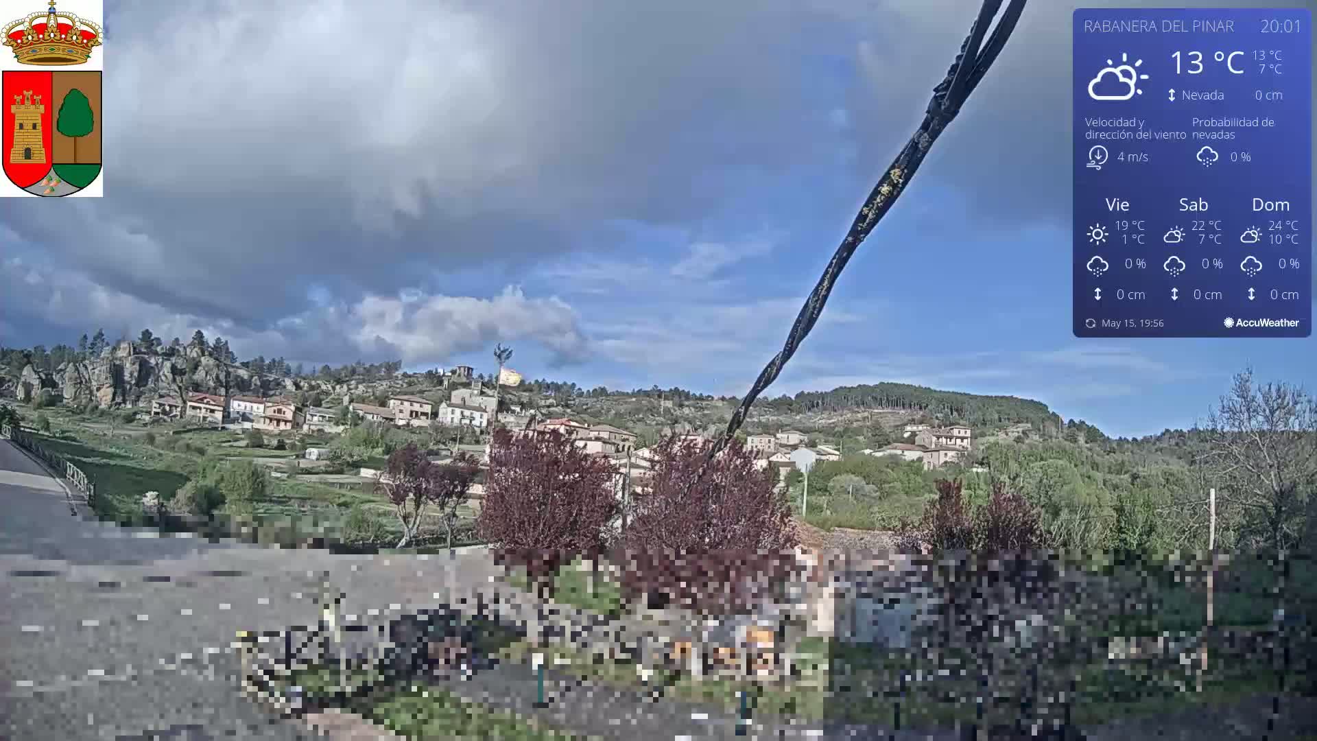 A partly cloudy day shows a small village nestled in a rocky hillside, with dark green trees and shrubs in the foreground.