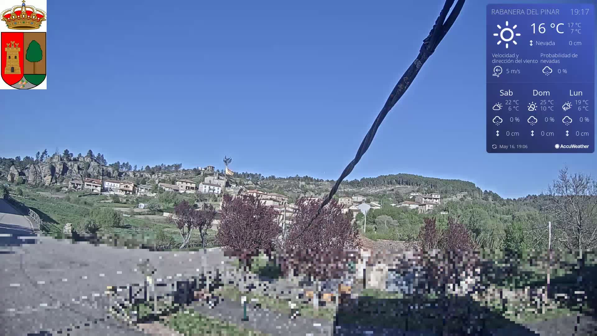 A small village nestled on a hillside under a clear blue sky is viewed from a slightly elevated vantage point on a sunny day with a gentle breeze.