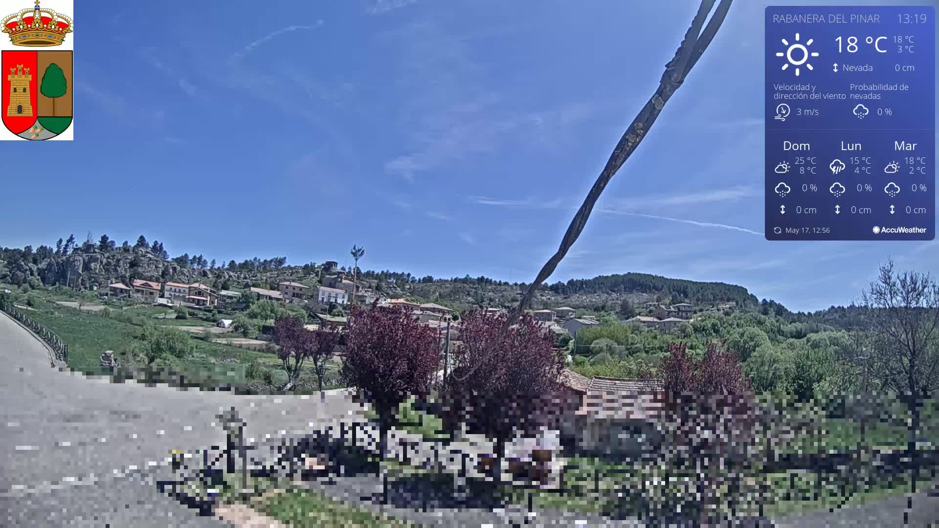 A mostly sunny, clear day reveals a small village nestled amongst green hills and rocky outcrops, with a partially visible road and dark-colored shrubs in the foreground.