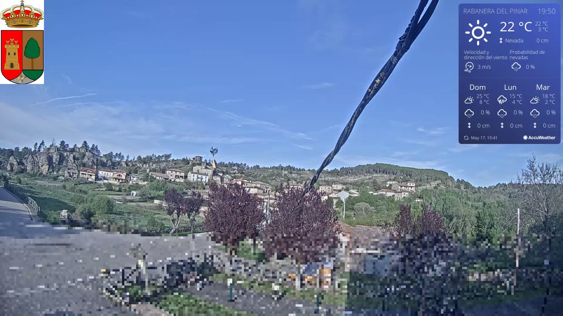 A small village nestled in a green valley under a clear blue sky is viewed from a slightly elevated position.
