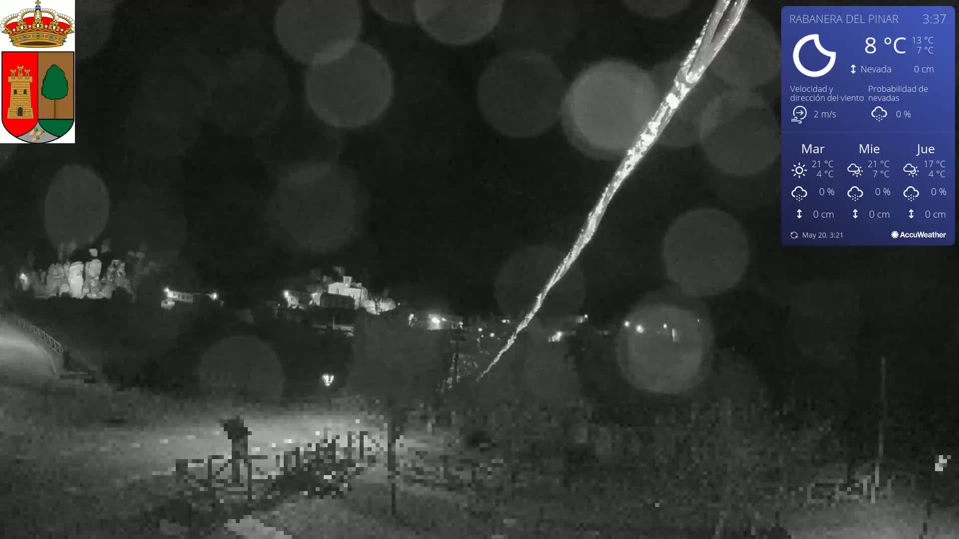 A nighttime view of a snow-covered landscape with a small town in the distance, under a dark sky with light rain or snow.