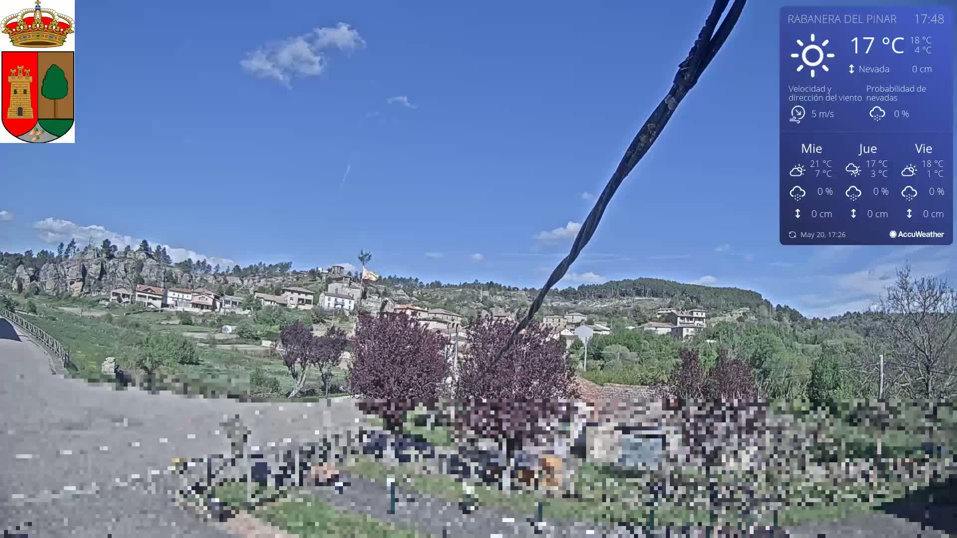A mostly sunny view of a village nestled in a hilly landscape with some trees and a road in the foreground, under a clear blue sky.