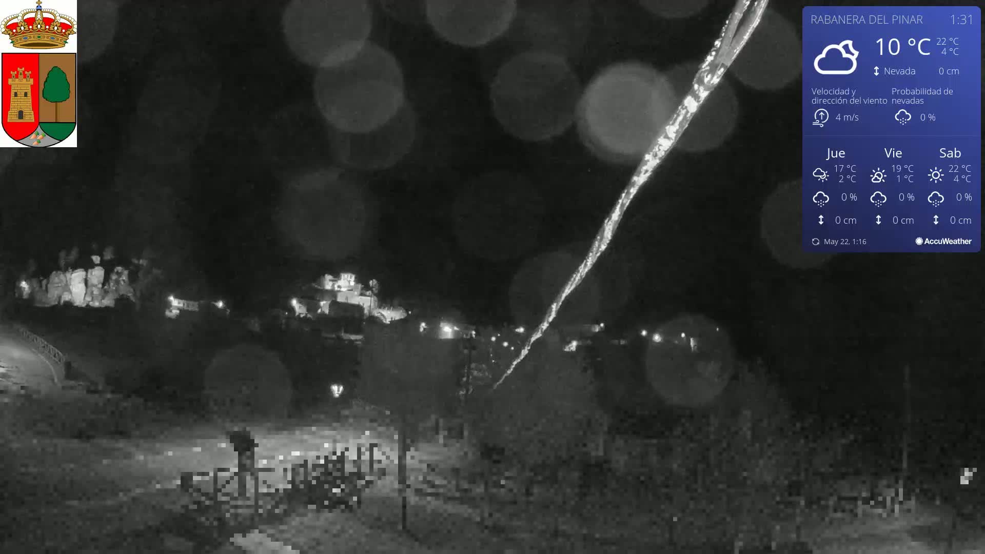 A nighttime, snowy scene shows a town illuminated by lights, with a long, thin streak of snow or ice visible in the foreground under a dark sky.