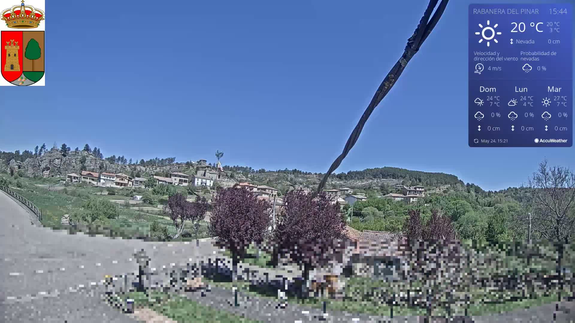 A small village nestled on a hillside under a clear blue sky is viewed from a slightly elevated position, with a road leading towards it.