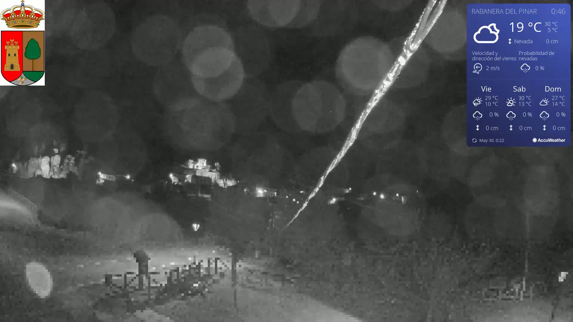 A nighttime, low-light view of a snow-covered landscape with a small town or village in the distance, under partly cloudy skies.