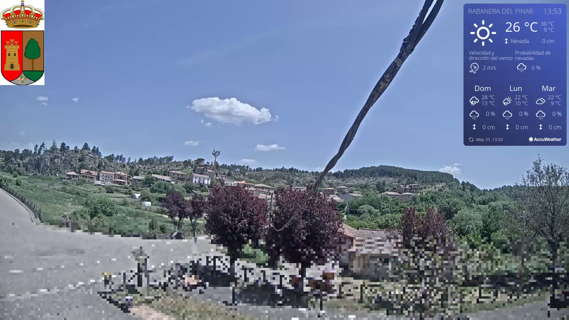 A partly sunny day shows a small village nestled in a hilly, green landscape with a few scattered clouds.