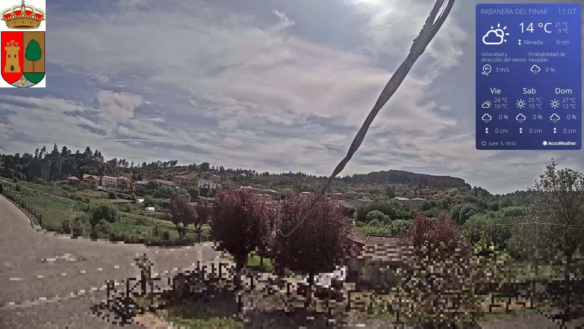 A partly cloudy day reveals a view of a small town nestled in a green hillside, with a long, dark object stretching diagonally across the foreground.