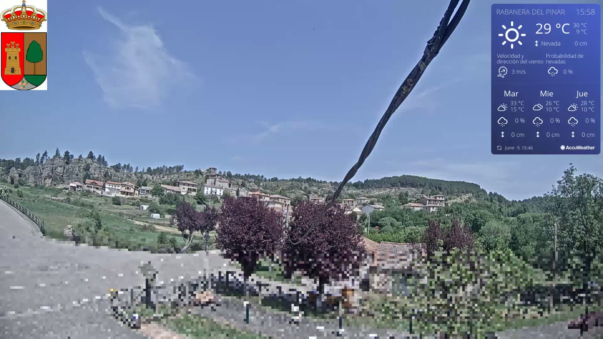 A village nestled on a hillside under a mostly clear, sunny sky is viewed from a road, with dark-colored shrubs in the foreground.